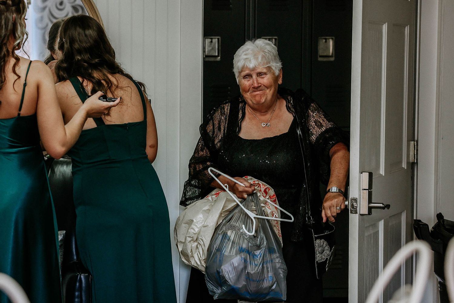 Elderly woman smiling while arriving at a wedding holding dress hangers and bags, greeted by bridesmaids in green gowns.