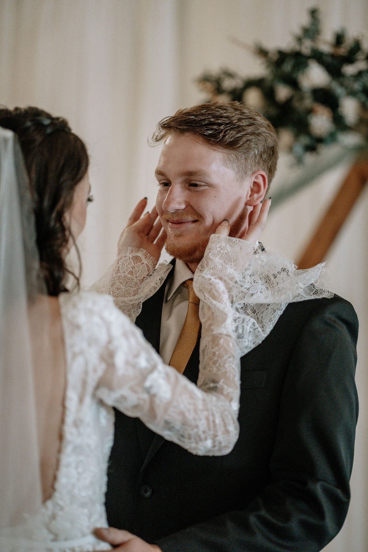 Bride in lace gloves tenderly holds groom's face during wedding ceremony, groom smiling warmly in dark suit.