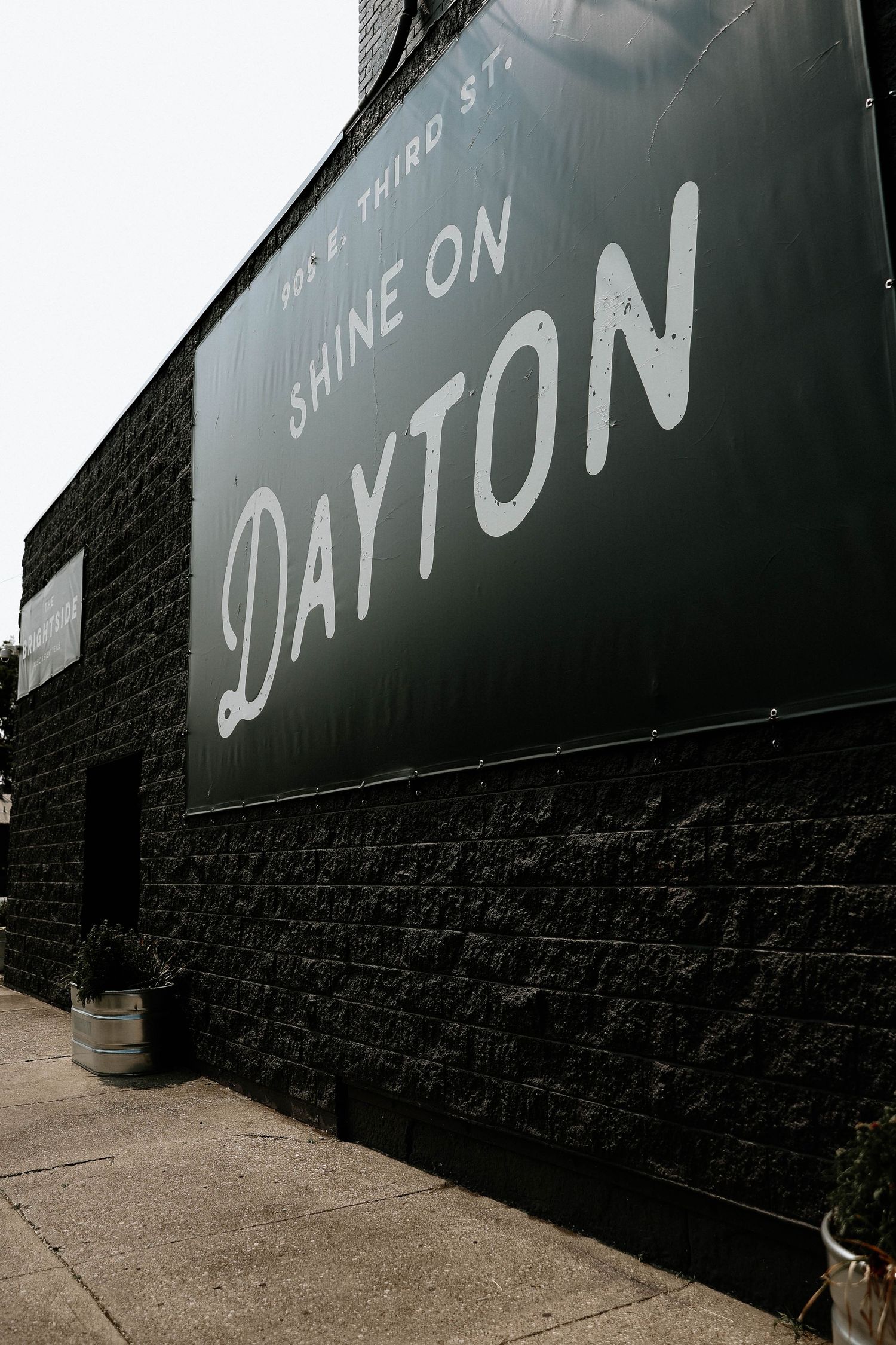 Black brick building exterior featuring a large green mural reading Shine On Dayton at 105 E Third St.