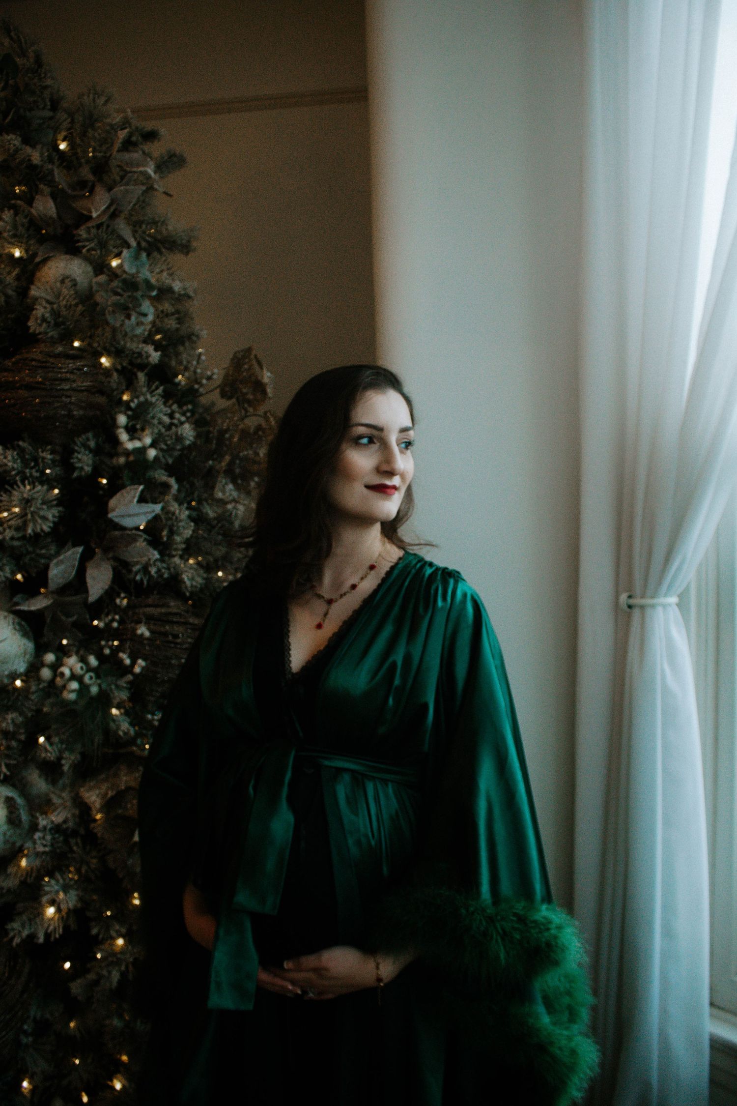 Pregnant woman in emerald green velvet dress by decorated Christmas tree at Mill Street Studios in Cincinnati, Ohio.