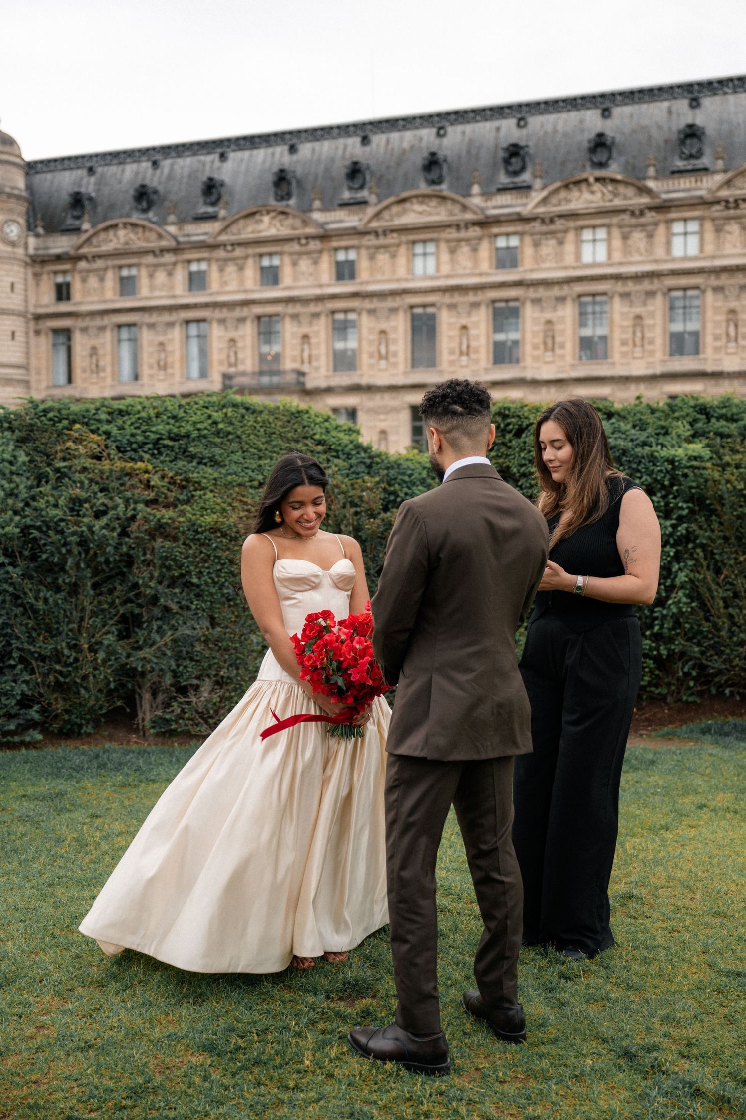 Bride and groom exchanging vows outdoors near the Louvre with red bouquet.