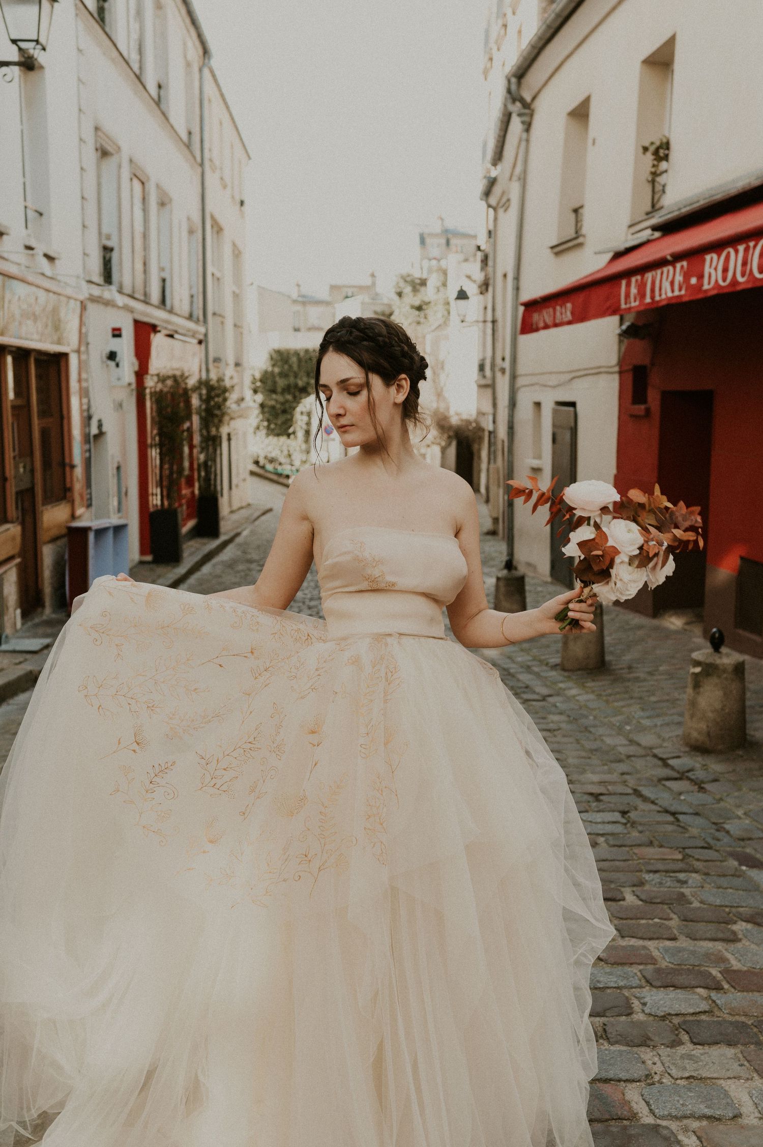 Bride in champagne tulle gown poses elegantly on historic European street with red storefront.