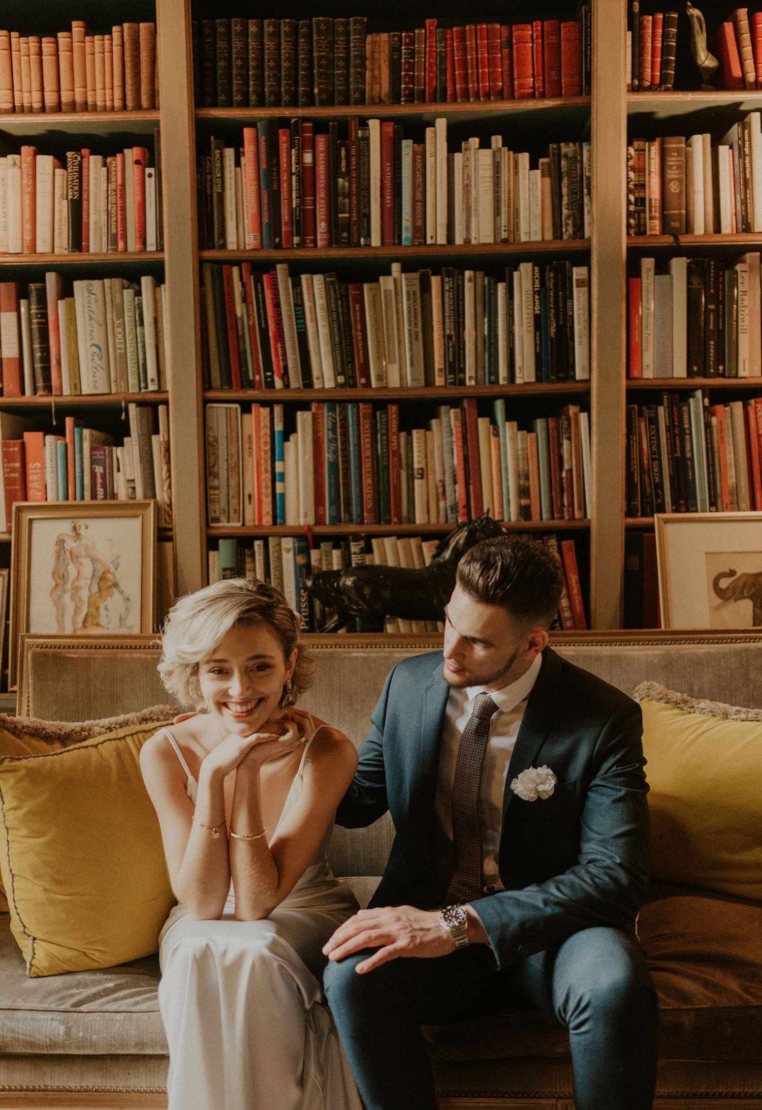 Intimate wedding portrait on yellow couch against wall of vintage books in library.