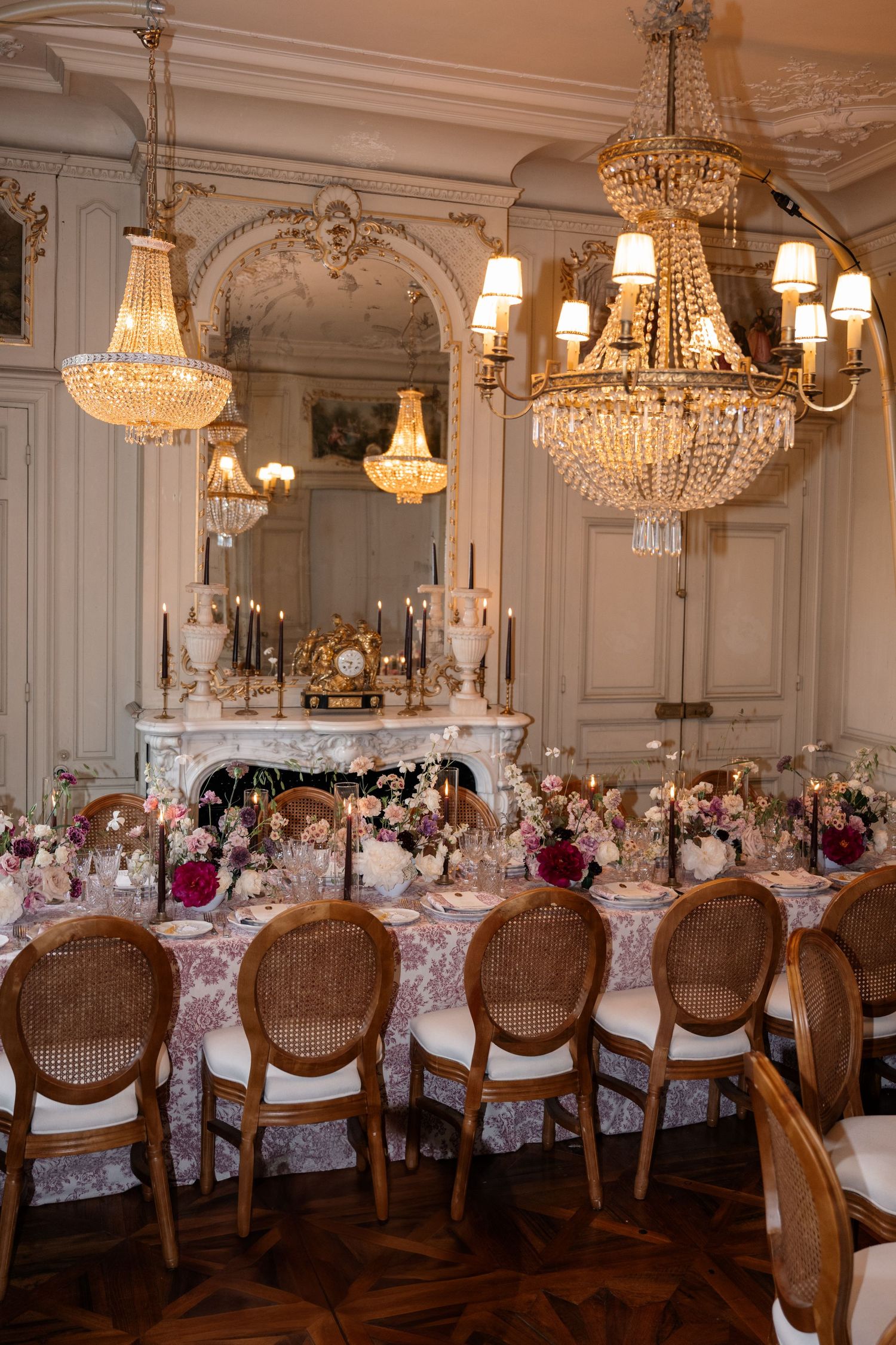Elegant dining room with crystal chandeliers, ornate fireplace and table set for formal dinner with floral arrangements.