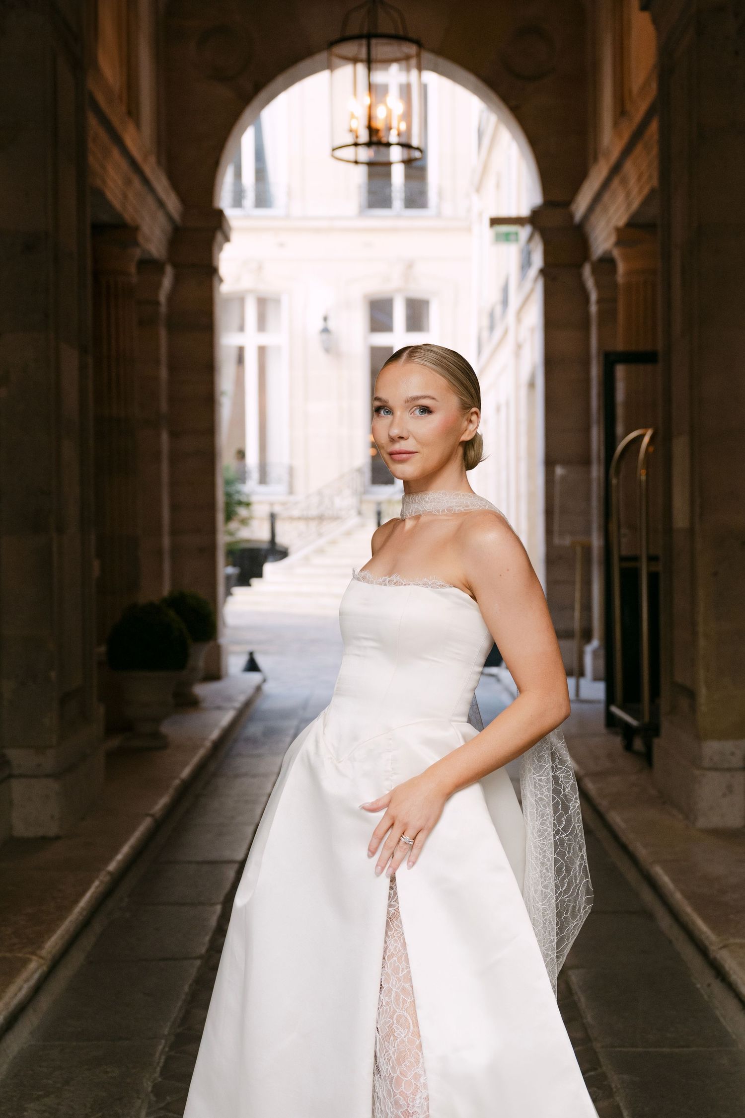 A bride in a white wedding dress poses elegantly in a grand stone archway corridor.