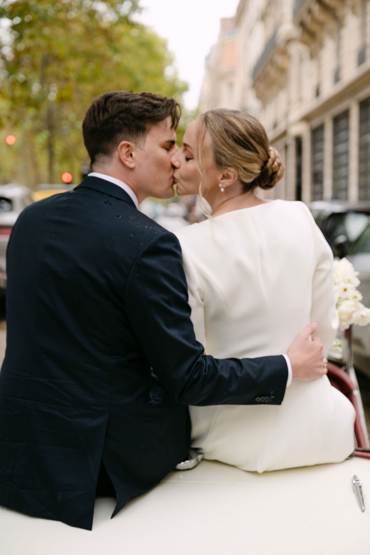 A tender moment shared between newlyweds in a classic car on a tree-lined Parisian street.