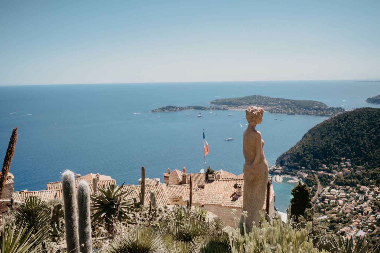 Mediterranean coastal view with stone statues overlooking a blue bay and distant peninsulas.