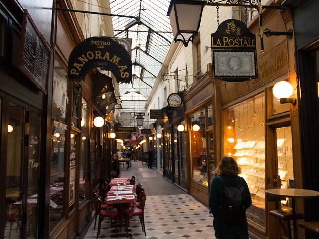 Elegant covered shopping arcade in Paris with ornate glass ceiling, vintage storefronts and warm lighting.