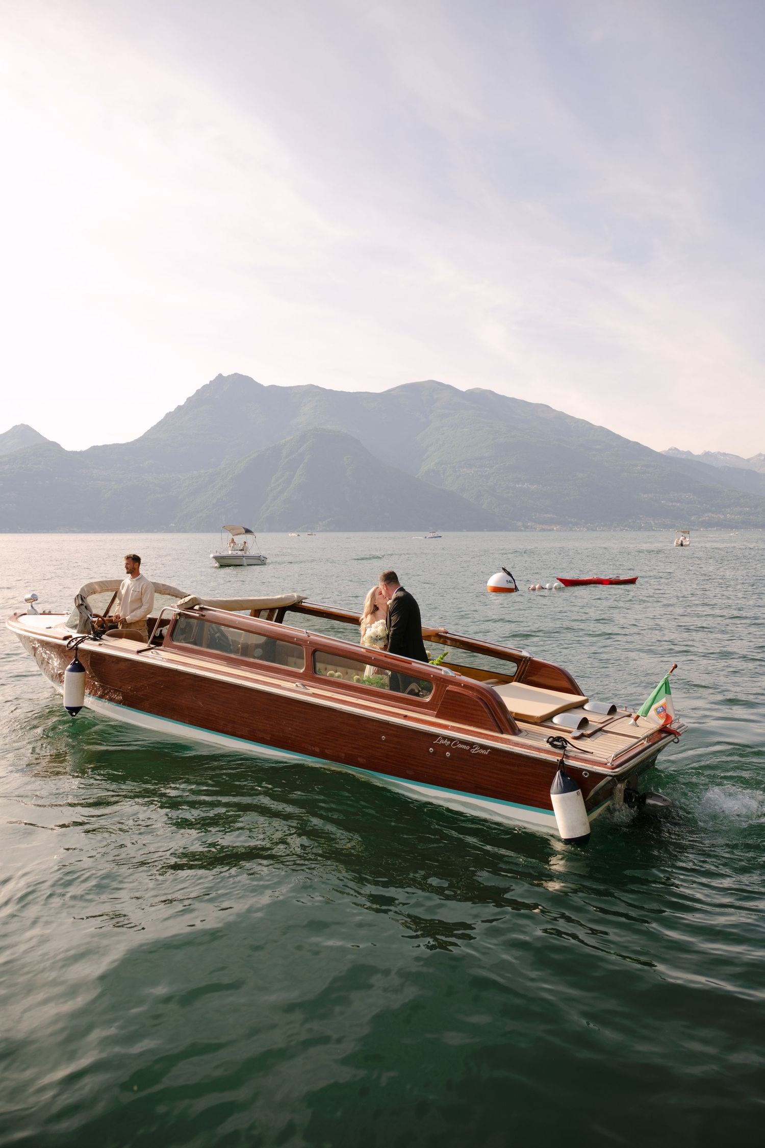 Couple kissing on a classic wooden boat on Lake Como with mountains in the background on a sunny day.