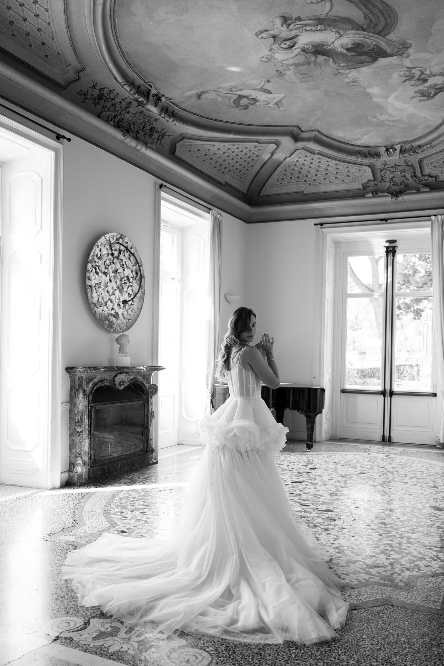 Bride in flowing white gown stands in elegant baroque room with ornate ceiling fresco and grand piano.