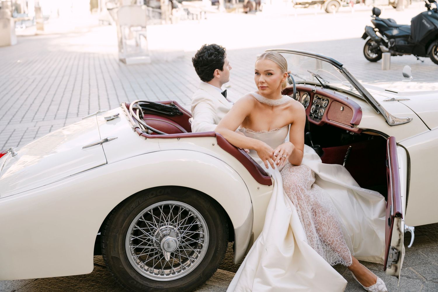 Bride in white gown sits in vintage convertible car with groom at wedding.