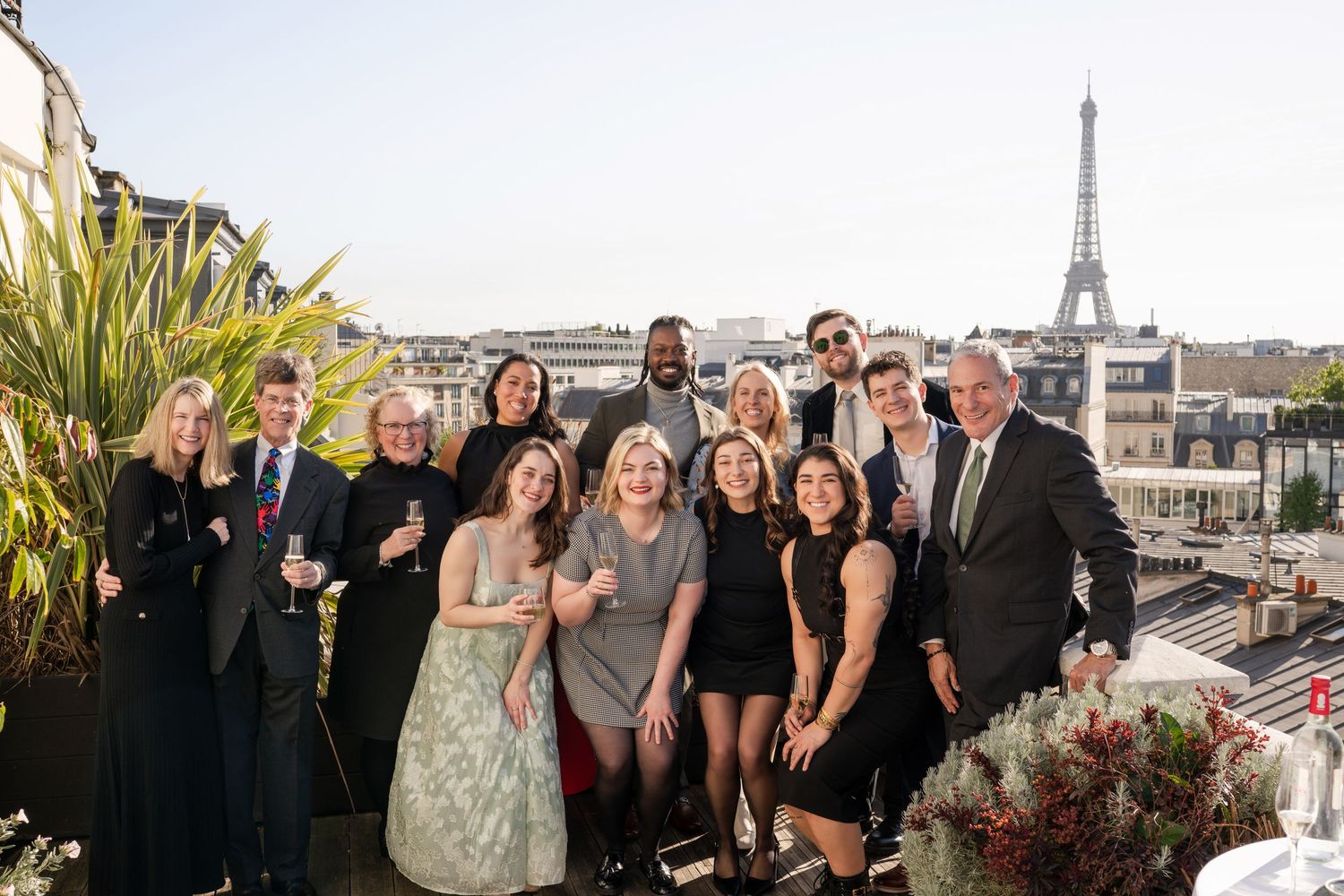 Group of elegantly dressed people posing on a Paris rooftop terrace with the Eiffel Tower visible in the background.