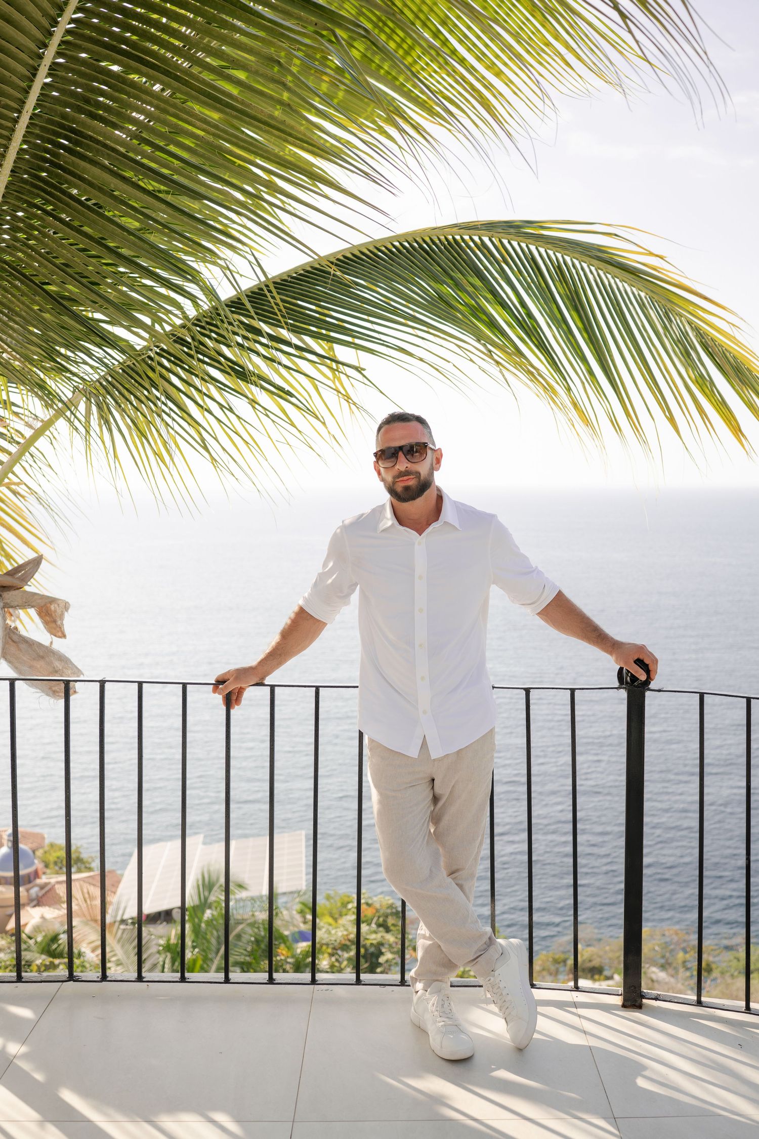 Man in white linen shirt and sunglasses standing on oceanview balcony with palm tree and blue sea backdrop.