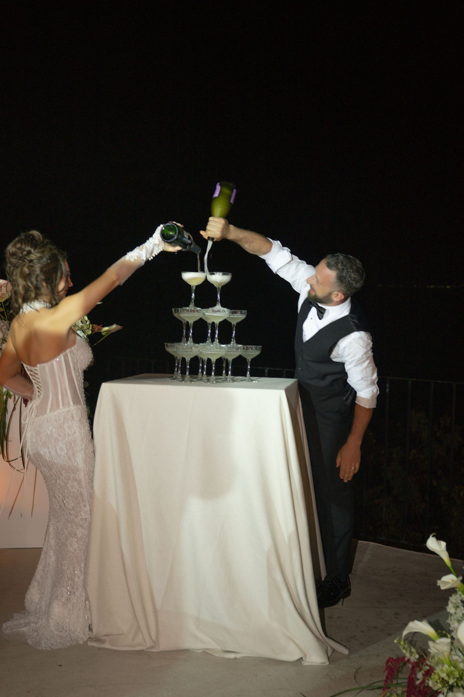 Bride and groom pouring champagne tower at wedding reception at night.