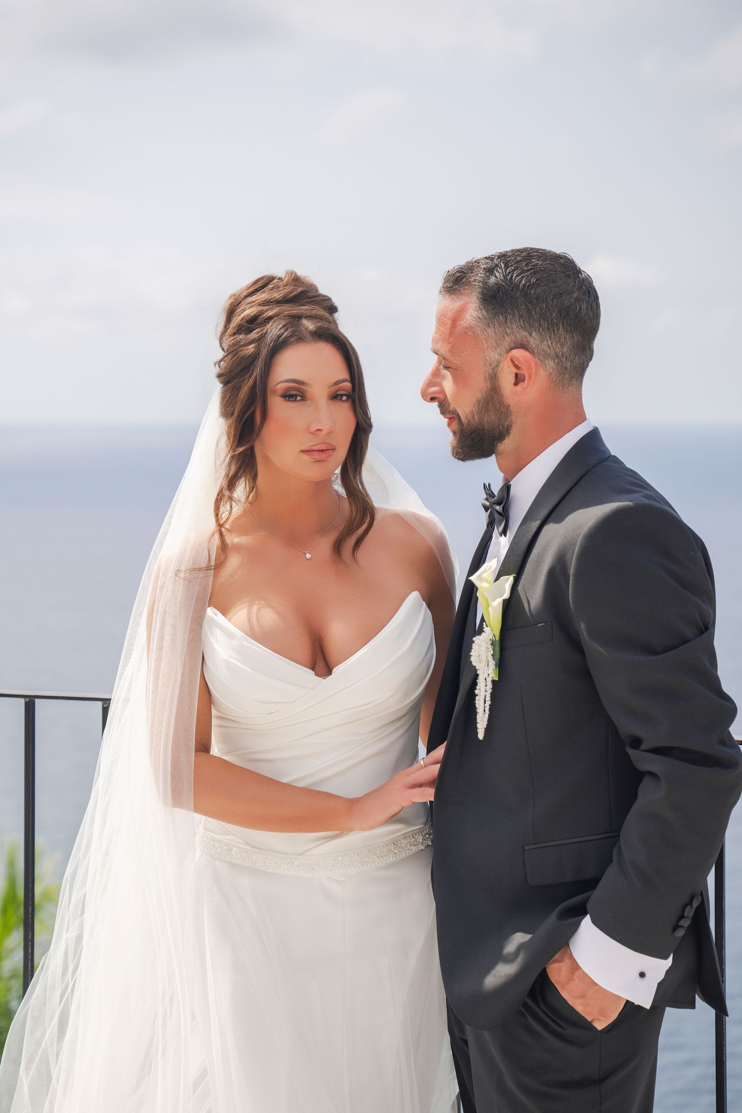 Bride and groom in elegant wedding attire sharing intimate moment on ocean-view balcony.