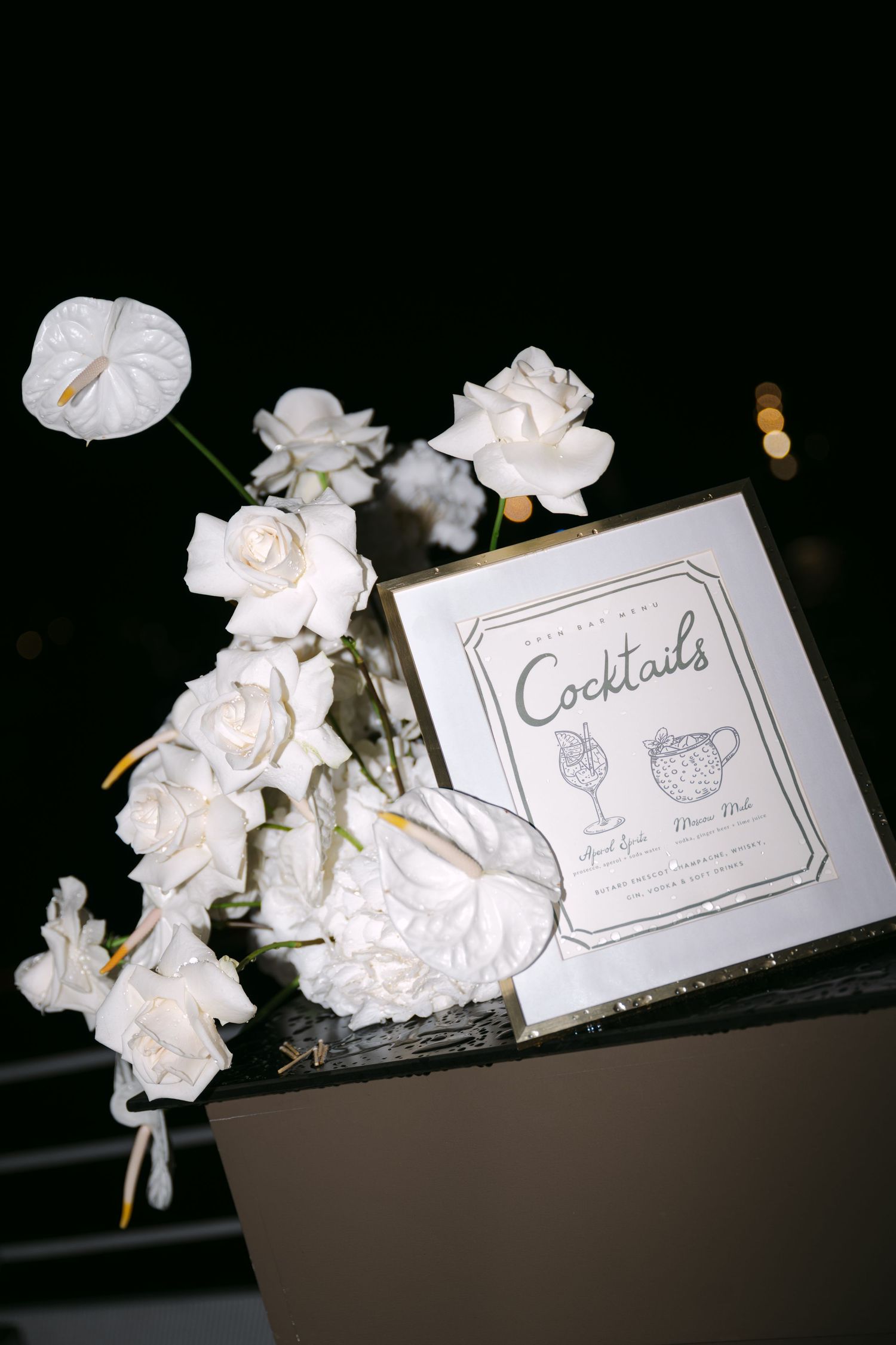 Elegant white floral arrangement with roses and anthuriums beside a cocktails menu sign at a wedding.