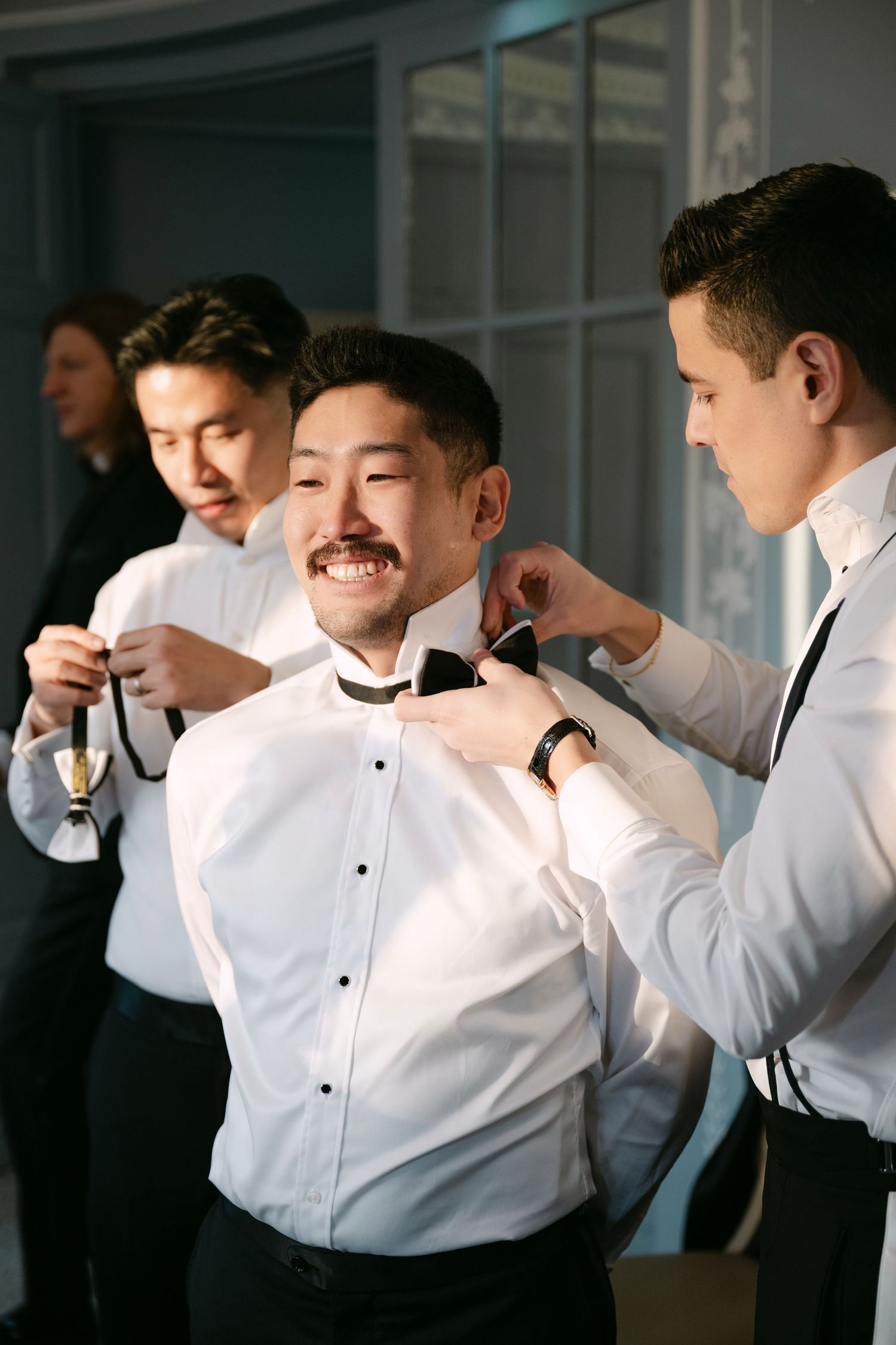 Groomsmen helping groom adjust black bow tie while getting ready for wedding ceremony.