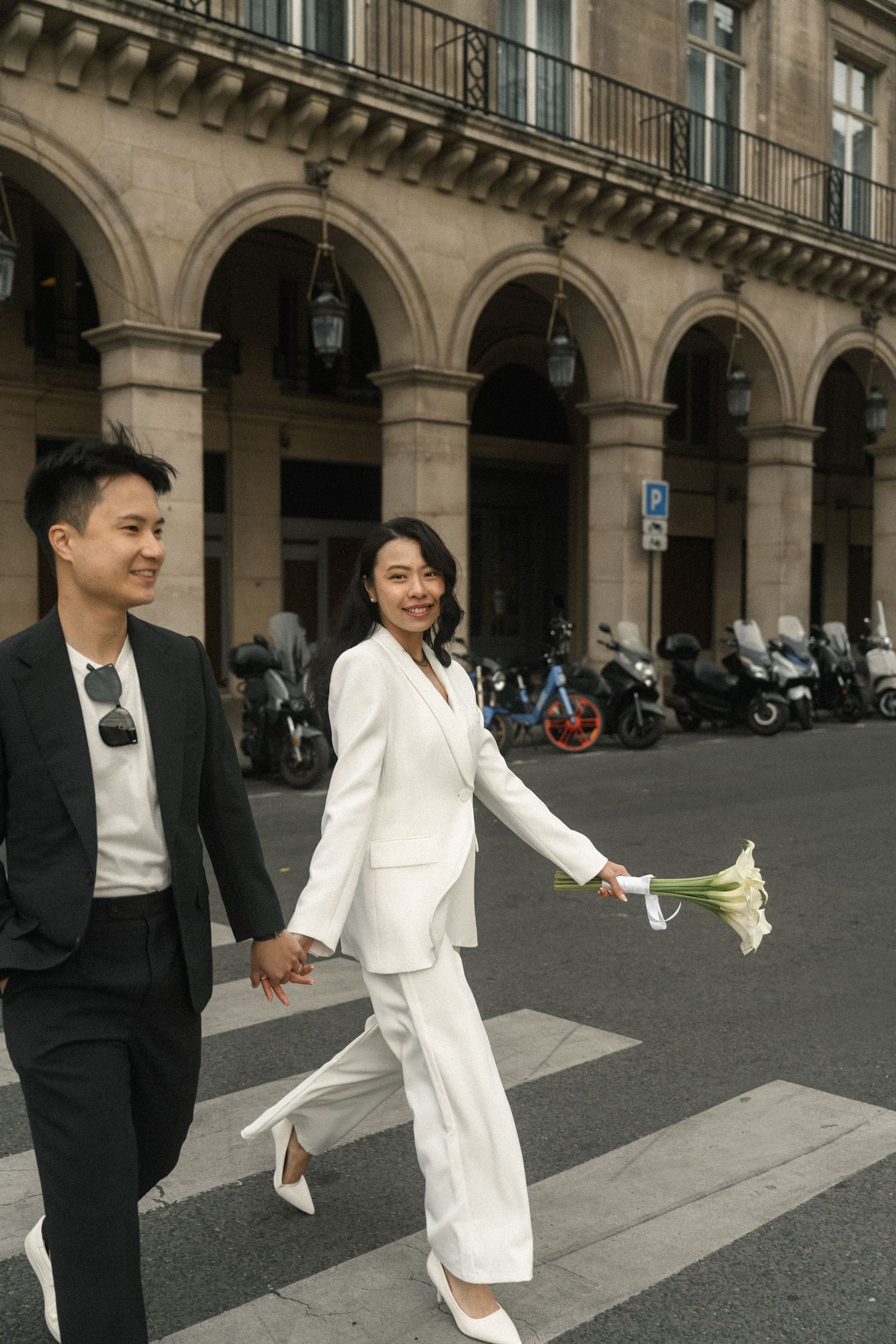 A smiling Asian couple in matching black and white suits cross a Paris street holding hands with flowers.