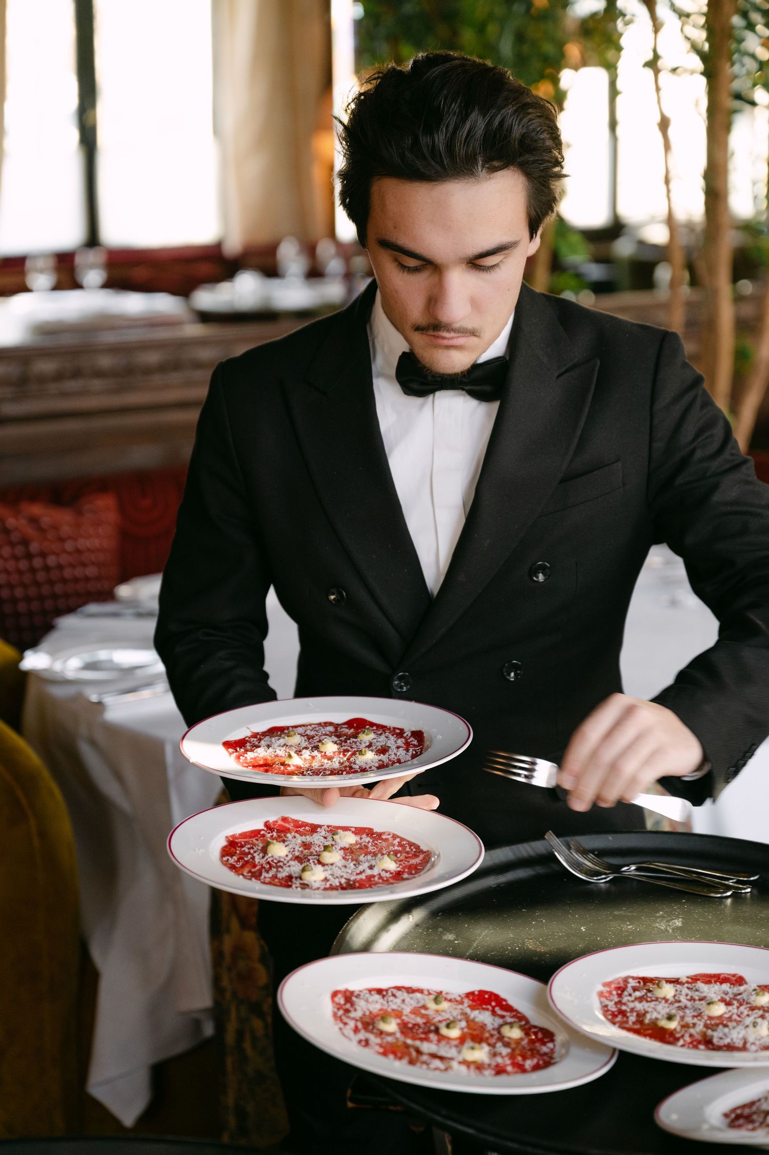 Elegant waiter in black tuxedo serving carpaccio dishes at upscale restaurant.