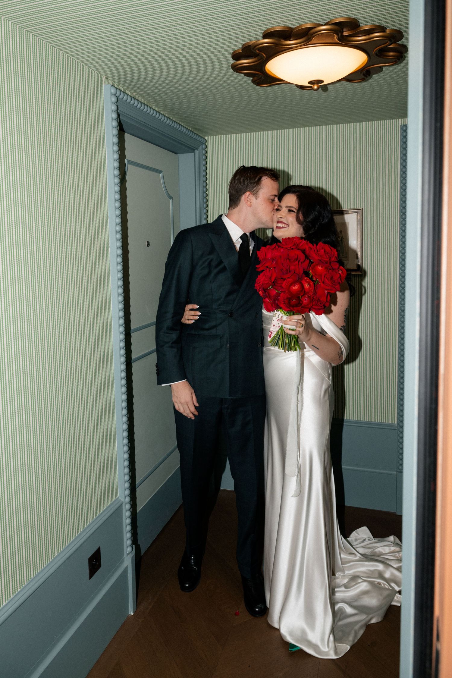 Groom in black suit kisses dark-haired bride holding red rose bouquet in a green wallpapered hotel corridor.