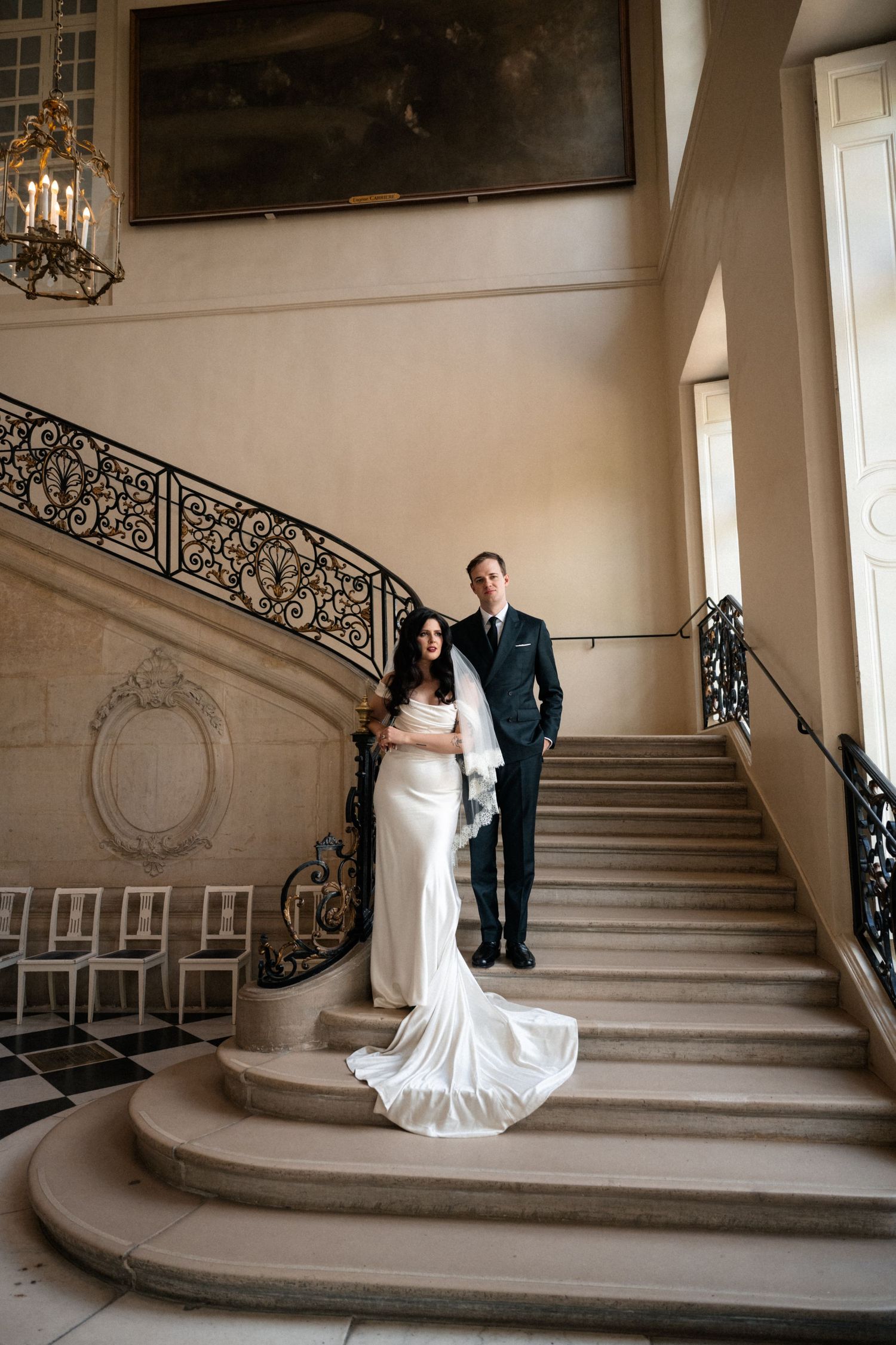 Bride and groom pose elegantly on sweeping stone staircase with ornate iron railings in grand French mansion.