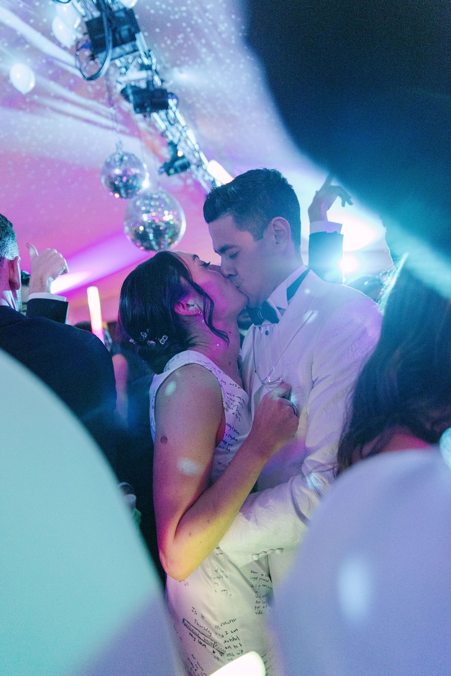 Bride and groom share passionate kiss on dance floor surrounded by guests at wedding celebration.