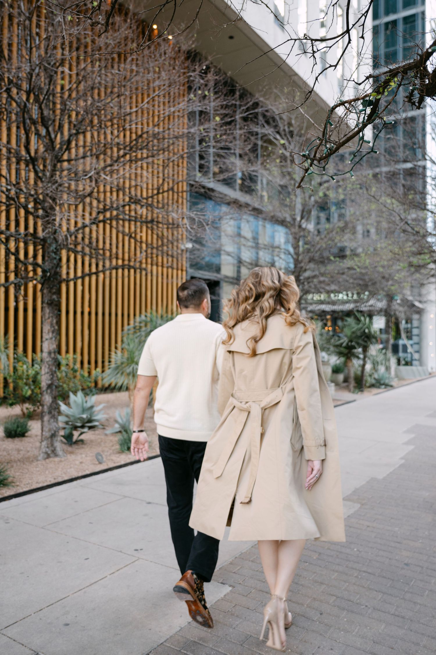 Stylish couple walking hand in hand past modern buildings, woman in beige trench coat and man in cream shirt.