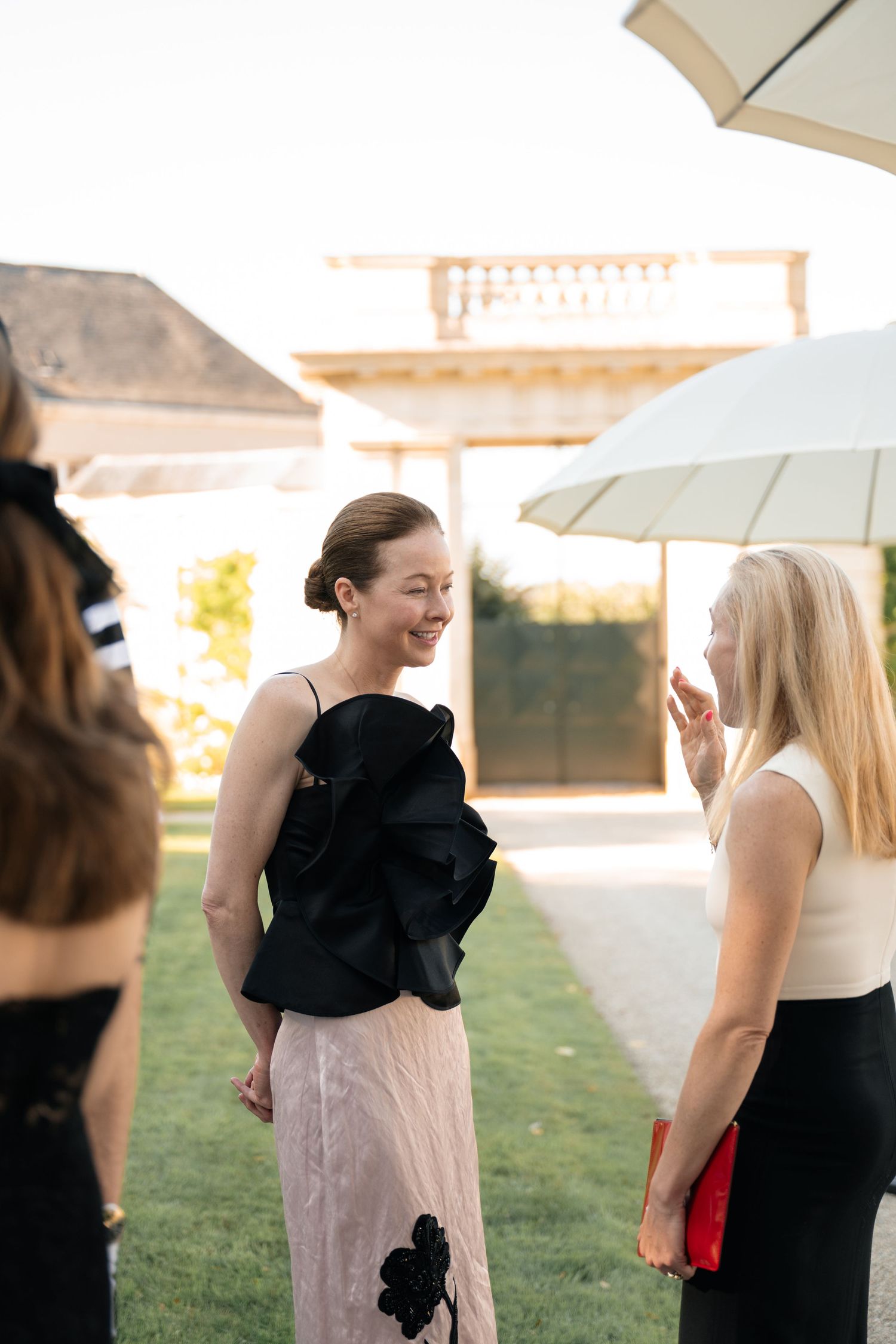 Two elegantly dressed women in black outfits conversing at an upscale outdoor garden event.