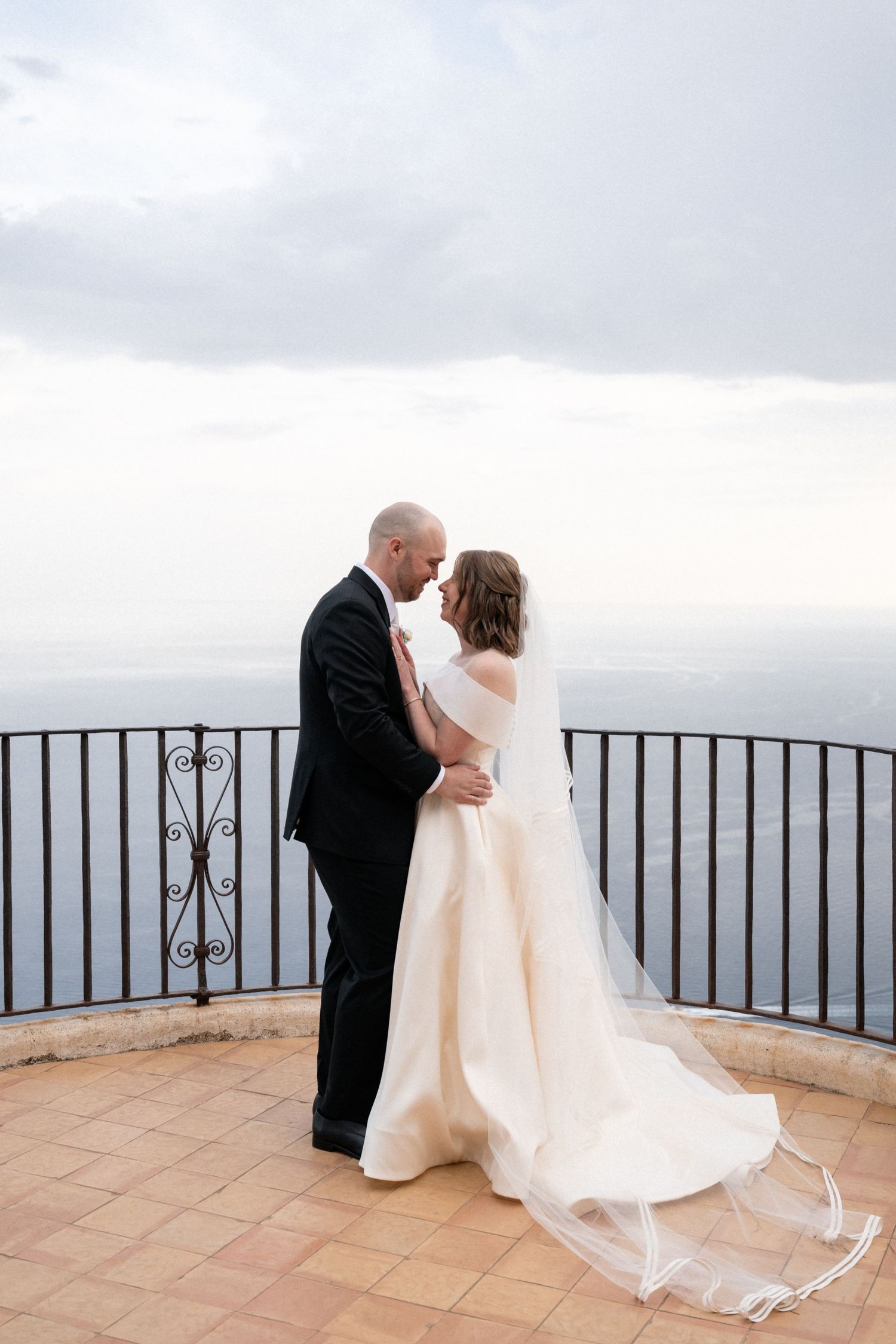 Bride and groom share a romantic embrace on a scenic balcony overlooking the sea on their wedding day.