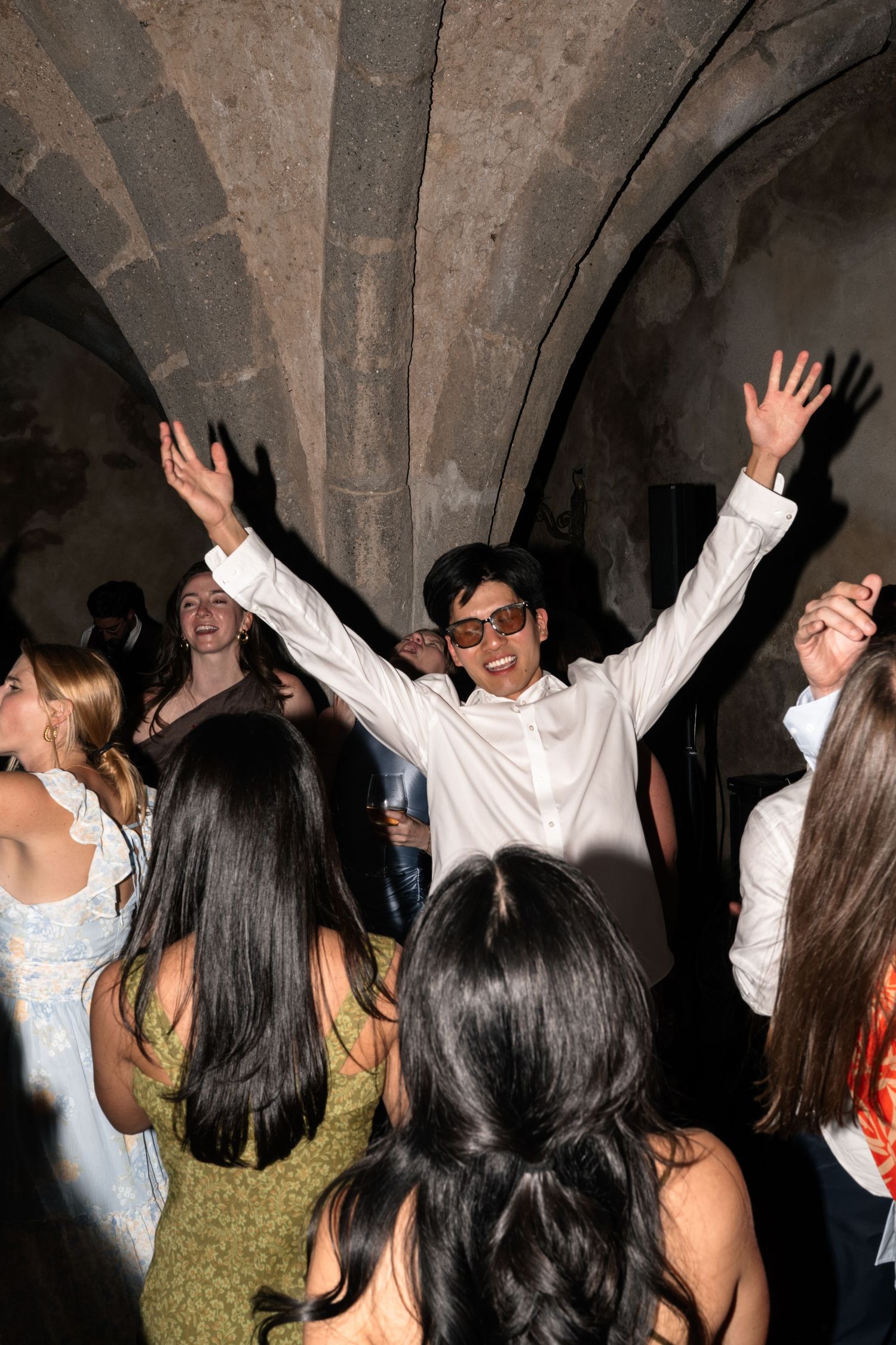 Joyful wedding guest in white shirt raises arms while dancing under stone arches at an Italian reception.