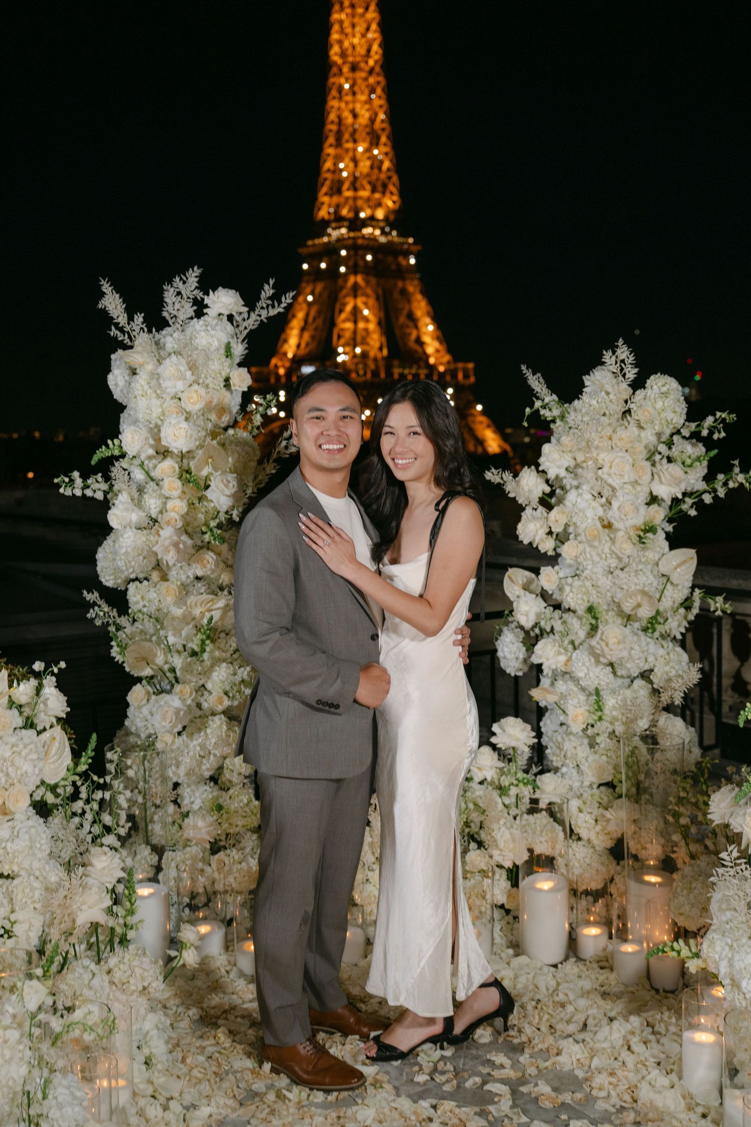 Happy engaged couple embraces among white flowers and candles with the sparkling Eiffel Tower behind them at night.