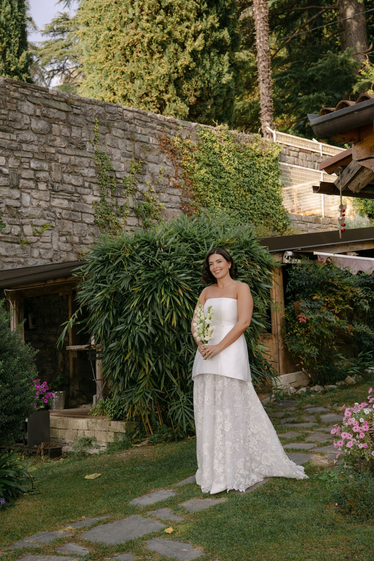 Bride in white strapless lace gown holding bouquet stands in lush garden with stone wall backdrop.