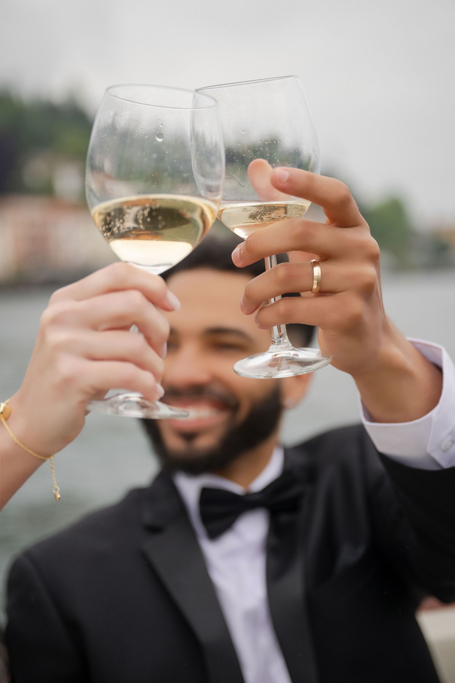 Newlywed couple toasting with white wine glasses outdoors, groom in black tuxedo with bow tie smiling near waterfront.