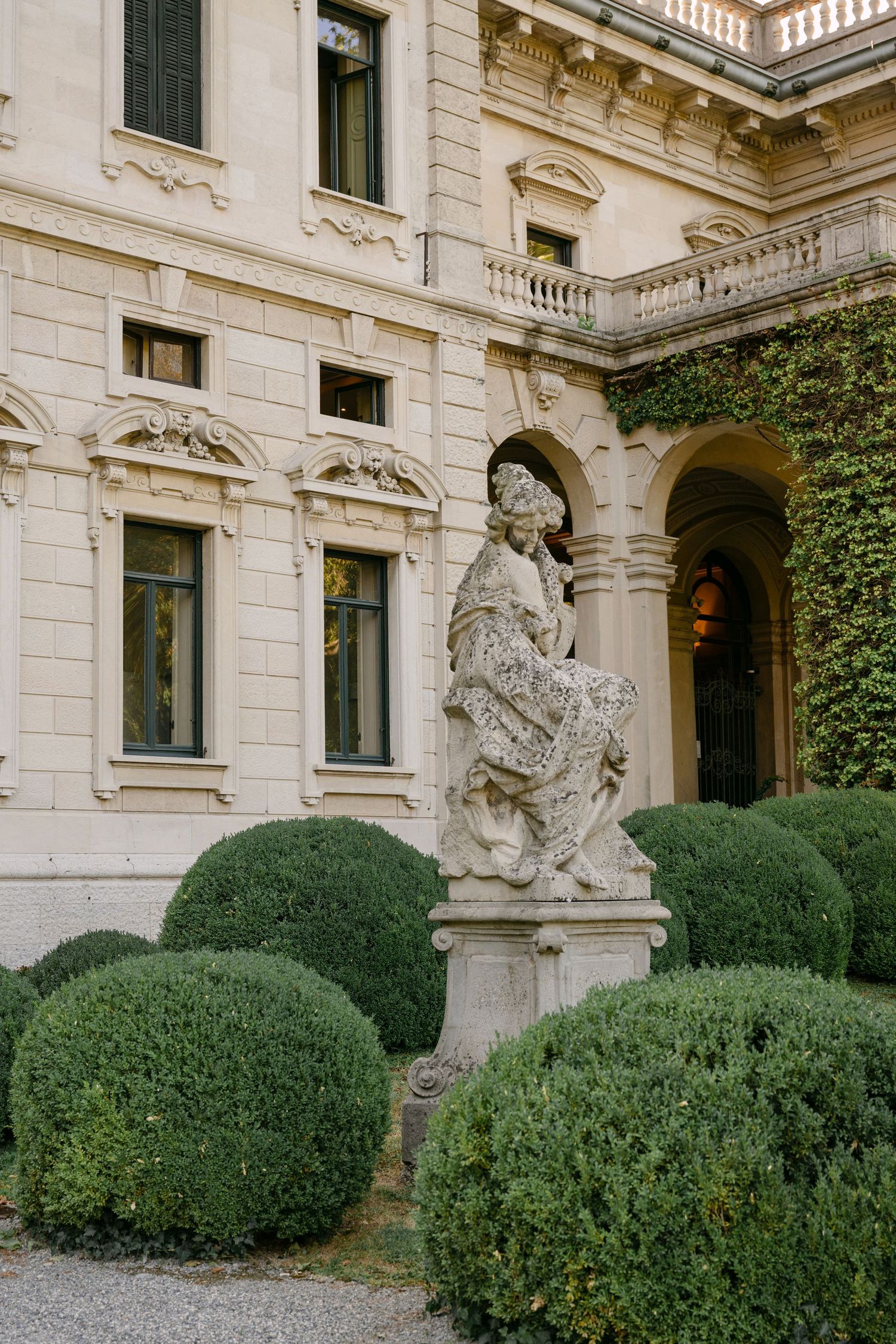 Baroque stone statue amid trimmed spherical hedges before an elegant Italian villa facade with arched colonnade.