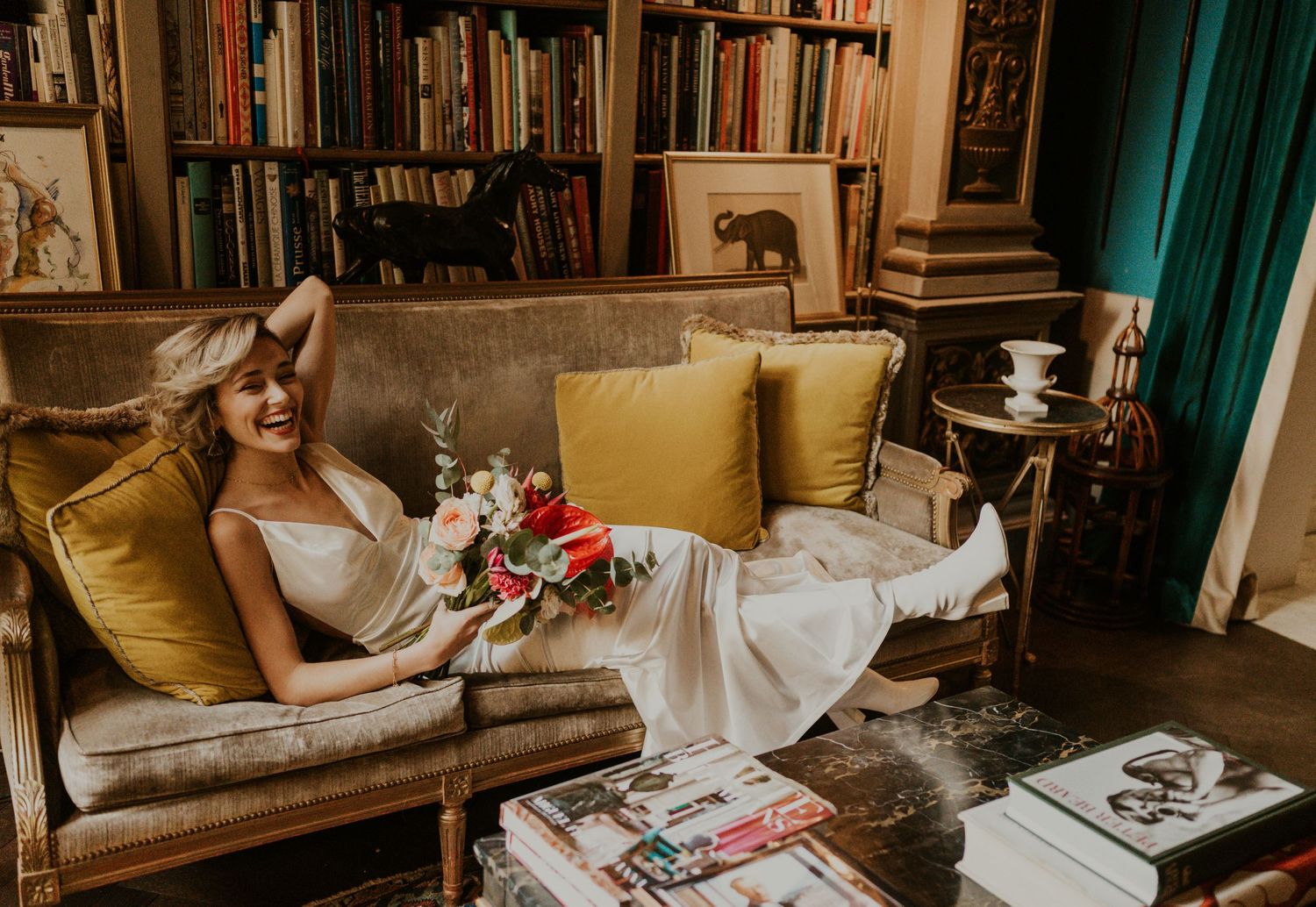 Bride relaxes on a vintage couch surrounded by bookshelves and yellow pillows.