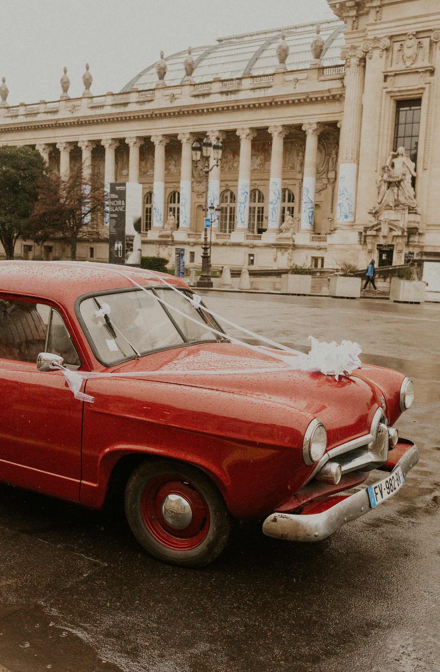 A vintage red car parked in front of a grand classical building with ornate columns.