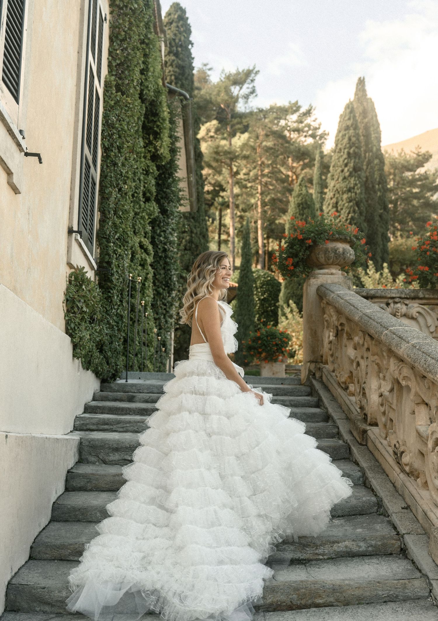 A flowing white wedding dress trails down stone steps alongside Italian cypress trees and an ornate balustrade.