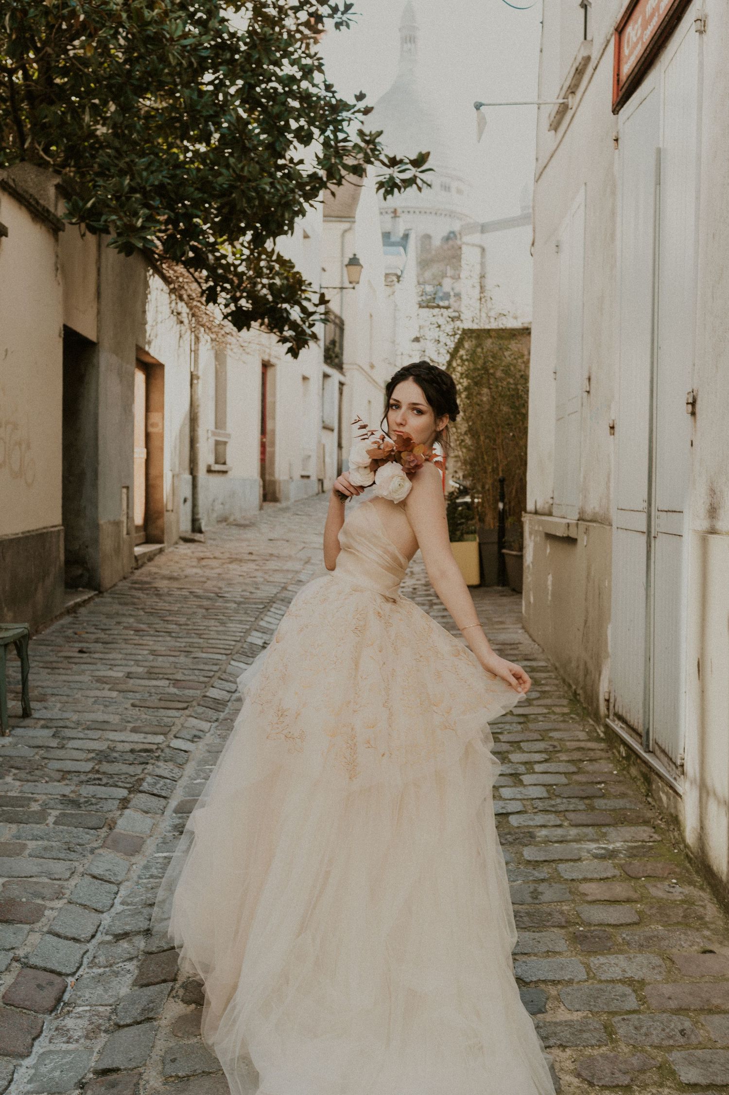 A bride in a strapless wedding dress poses playfully in a narrow European street with stone pavement.
