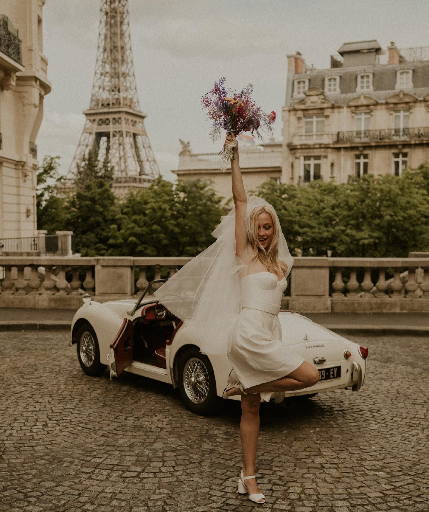 A white dress and vintage car create a romantic scene in front of the Eiffel Tower in Paris.