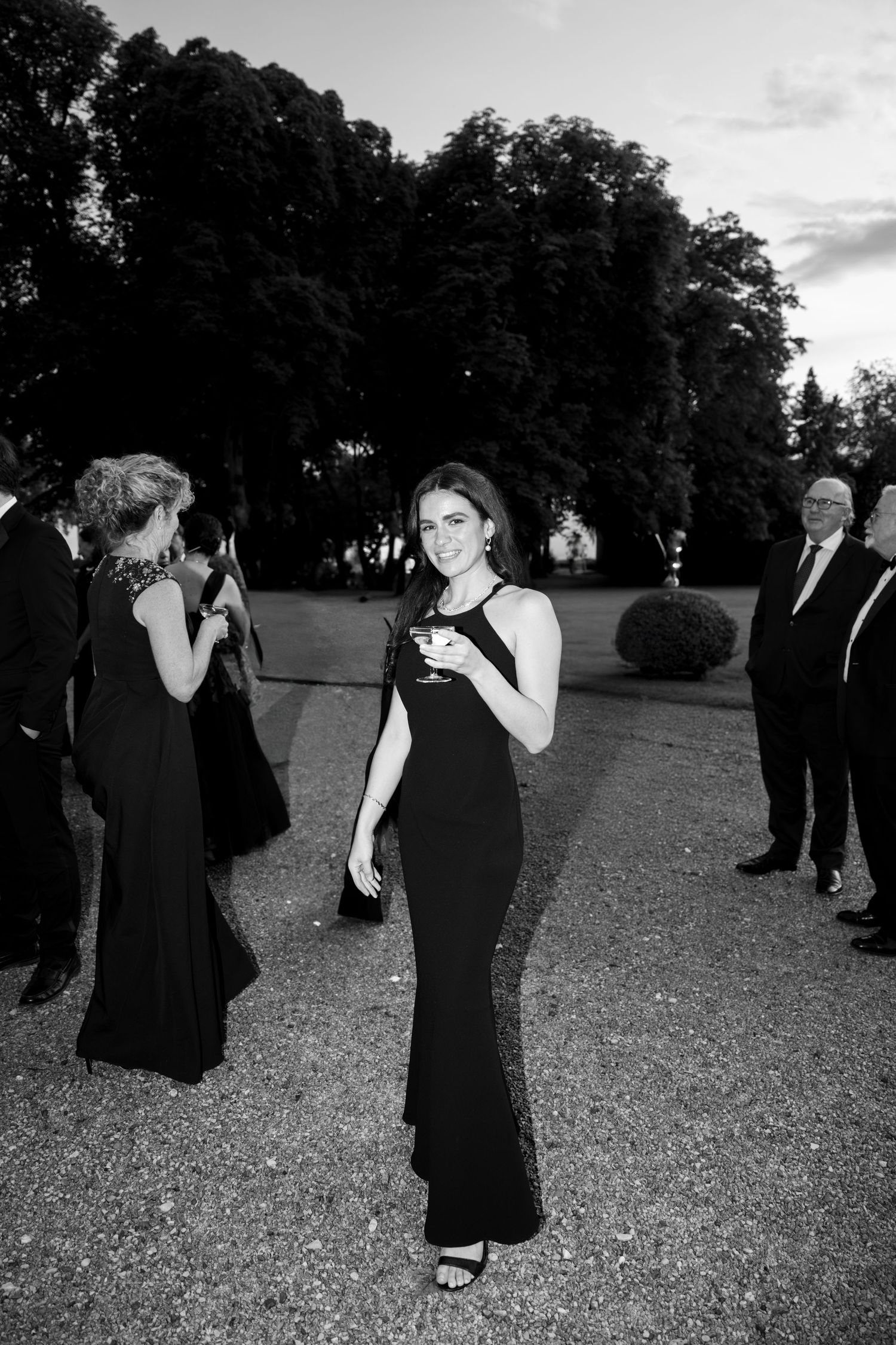 A group of people in formal black attire gathered outdoors at dusk under tall trees.