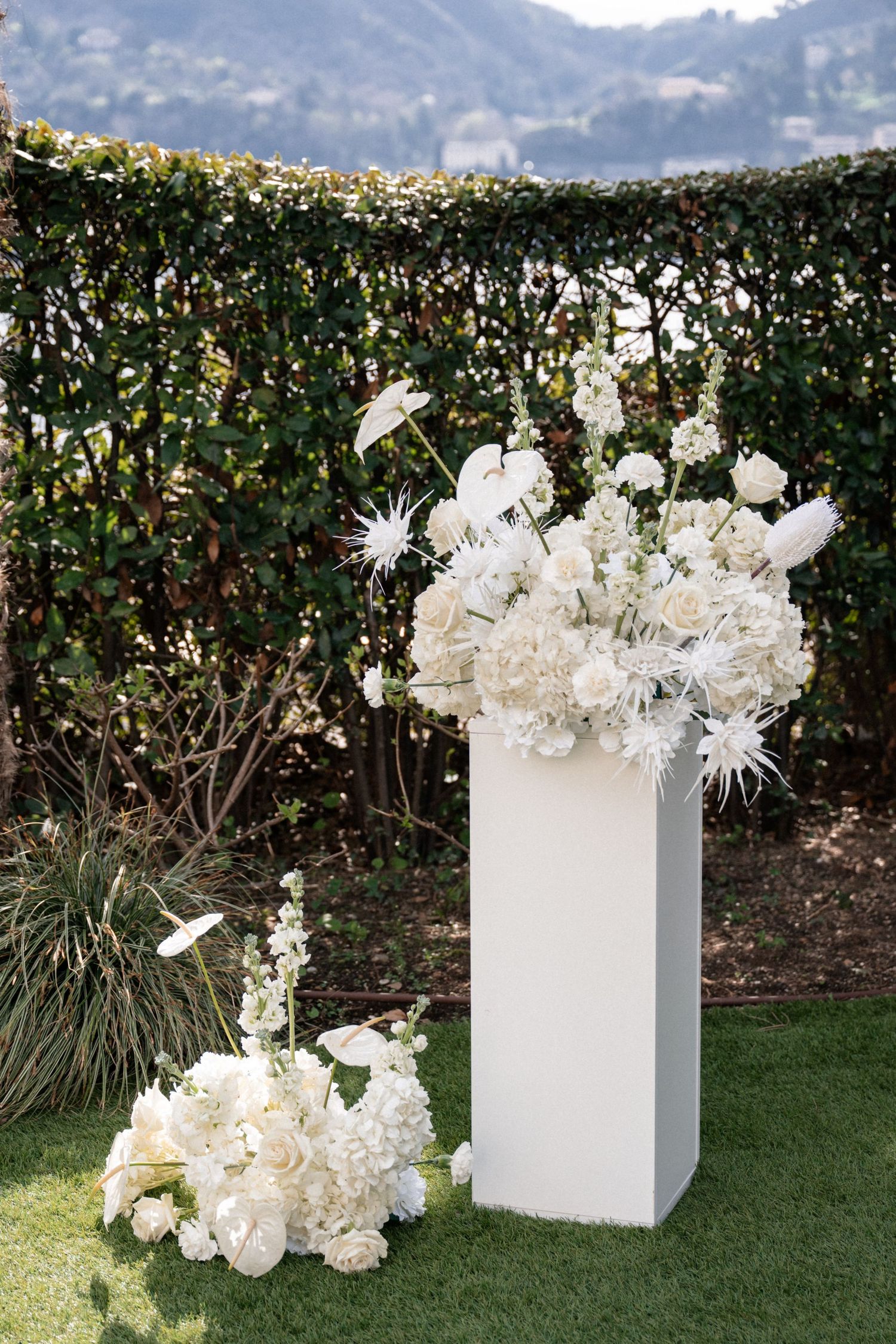 Lush all-white floral arrangement on a modern white pedestal at an outdoor wedding ceremony.