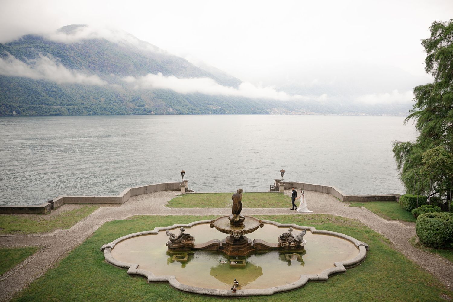 Wedding couple posing near ornate fountain in manicured garden overlooking misty lake and mountain landscape.