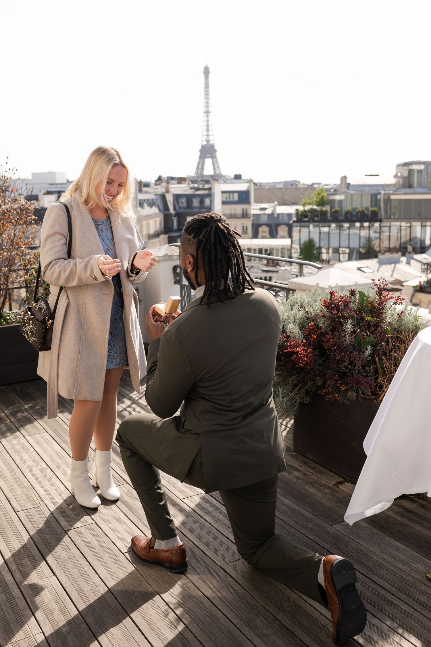Man proposing on one knee to woman on Parisian rooftop terrace with Eiffel Tower in background.