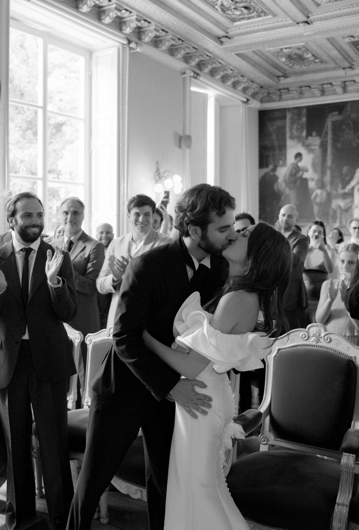 Black and white photo of a couple sharing their first kiss at a wedding ceremony with guests applauding in an elegant venue.