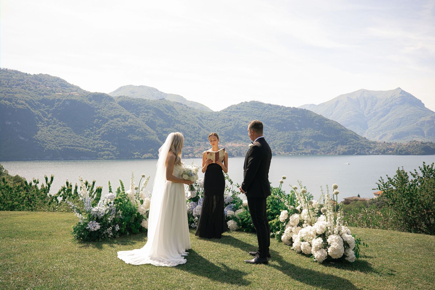 Outdoor wedding ceremony by a mountain lake with bride, groom, and officiant on green lawn with white floral arrangements.
