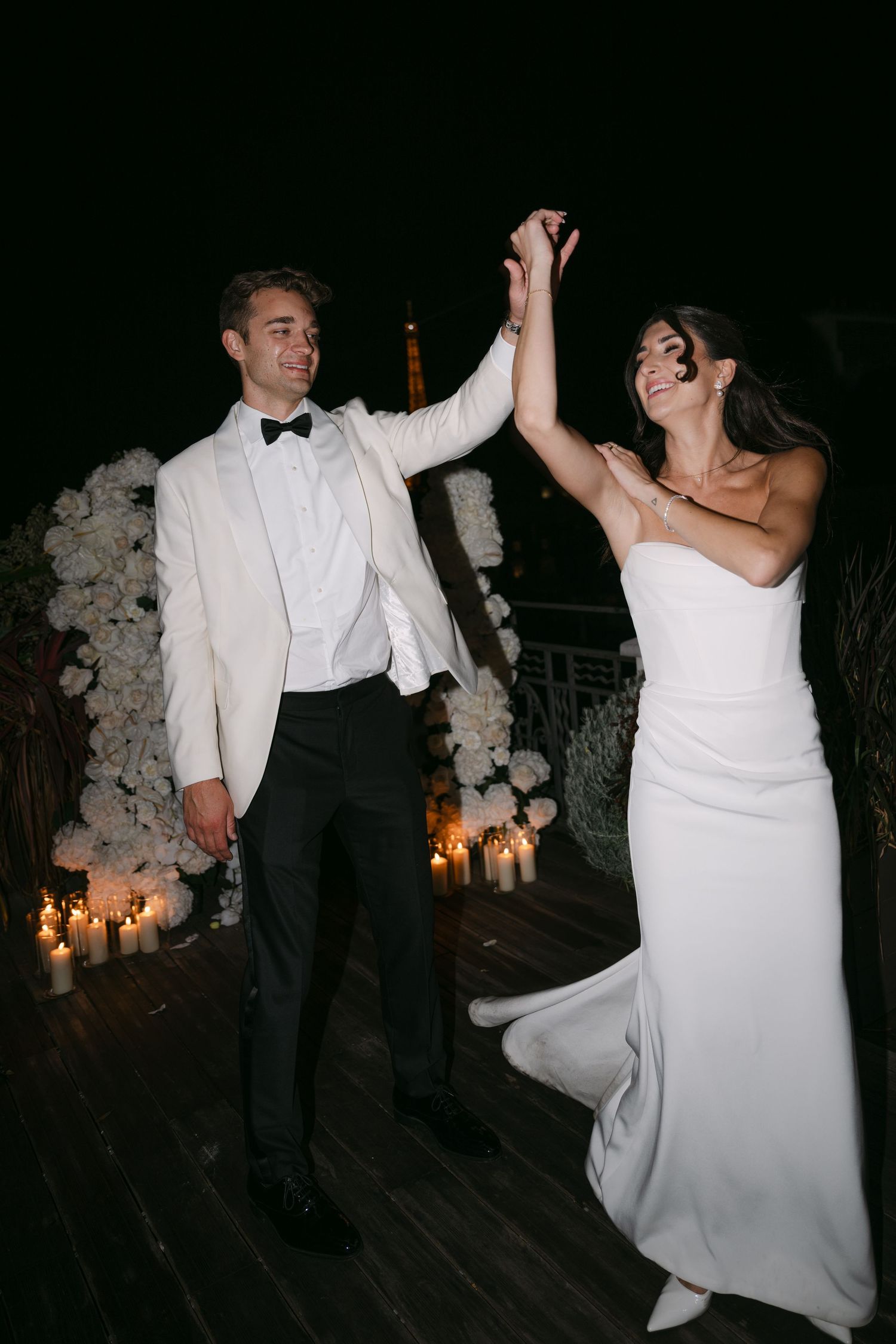 Bride and groom dancing joyfully on a candlelit rooftop with the Eiffel Tower at night.