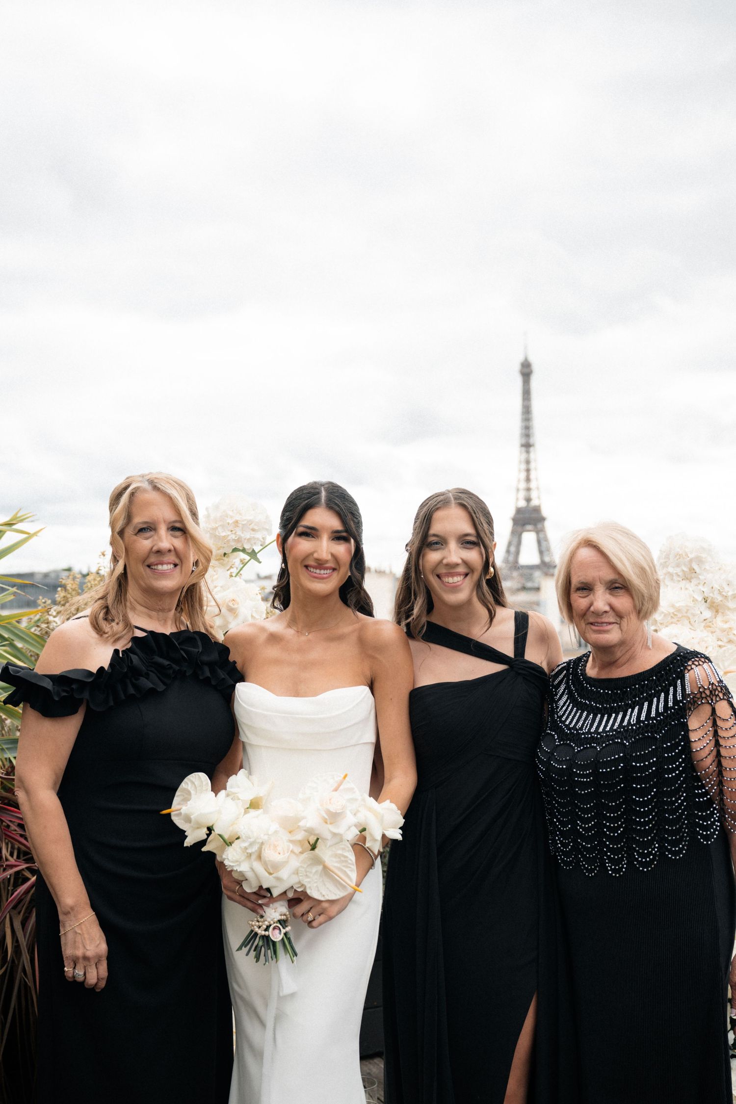 Bride holding white bouquet poses with three women in black dresses, Eiffel Tower visible in background.