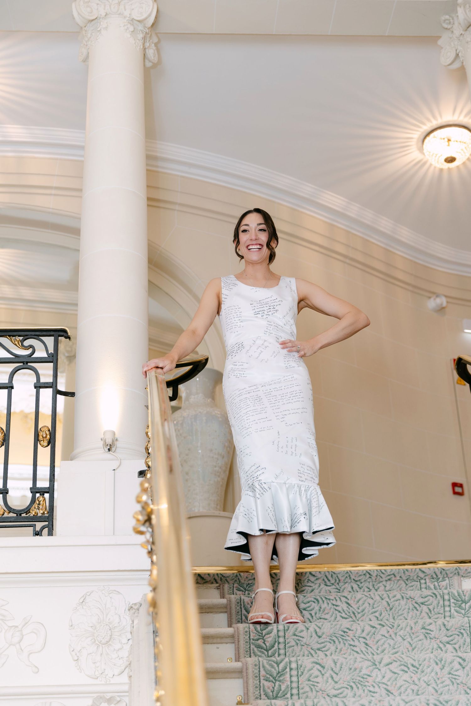 Elegant woman in white mermaid dress smiling on grand staircase with ornate columns.