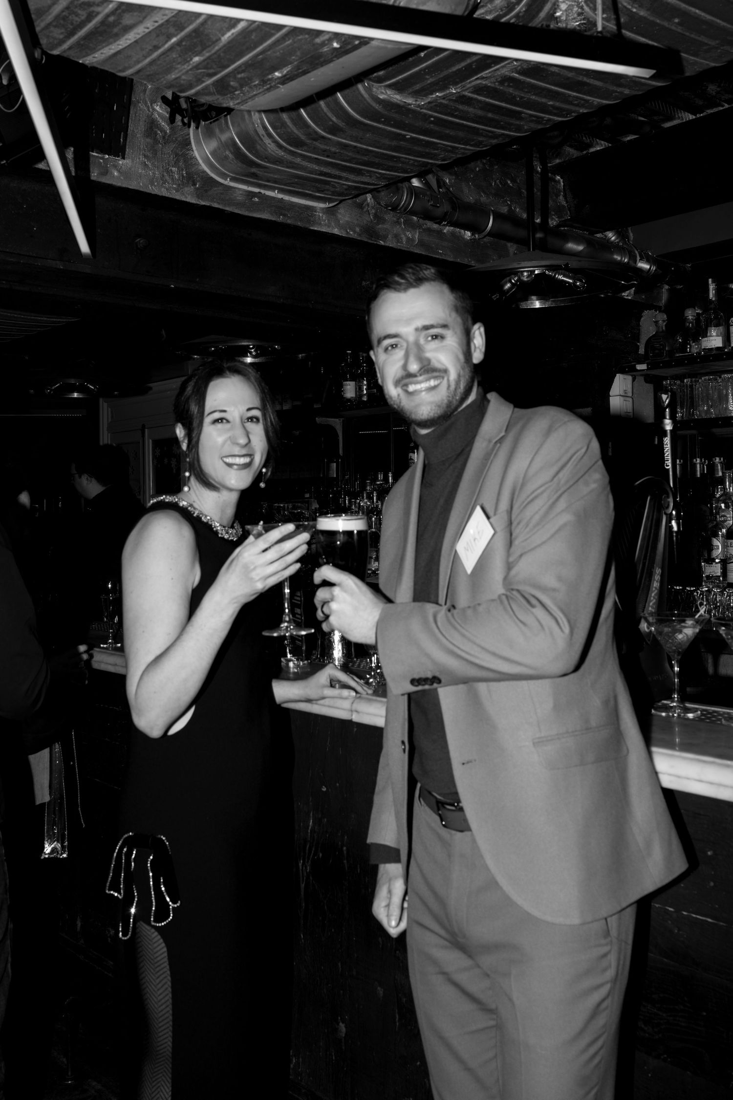 Black and white photo of smiling couple toasting with drinks at a bar.