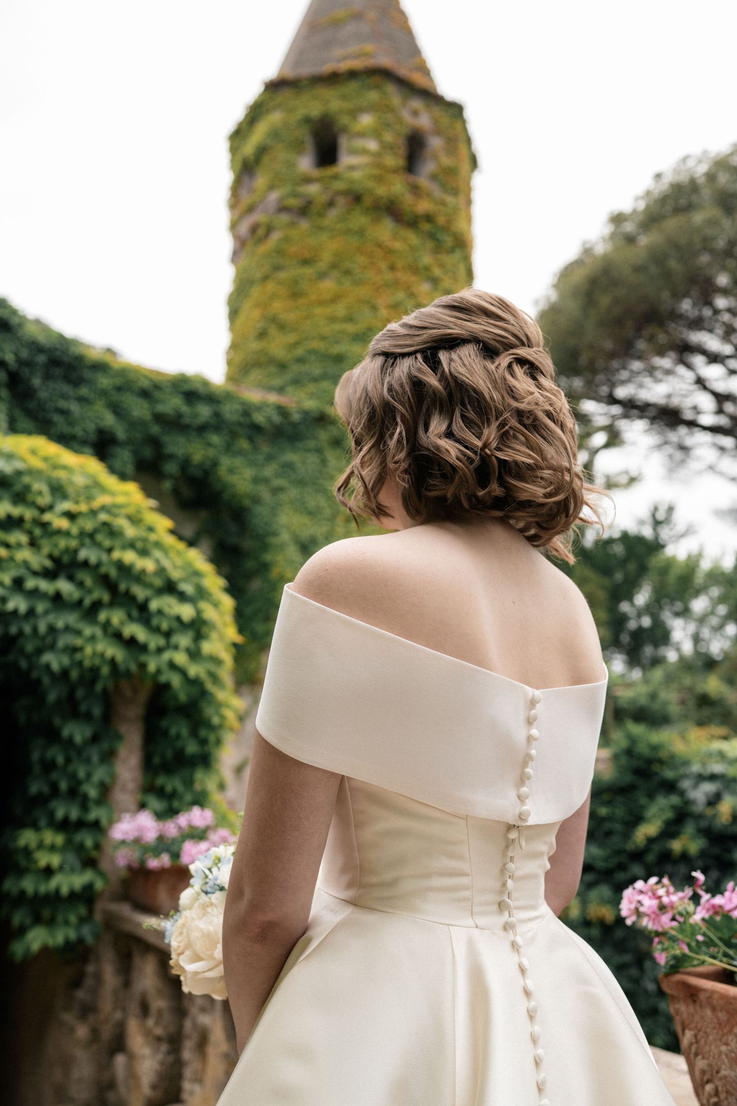 Bride in off-shoulder ivory wedding gown with button back detail stands before ivy-covered tower in garden.